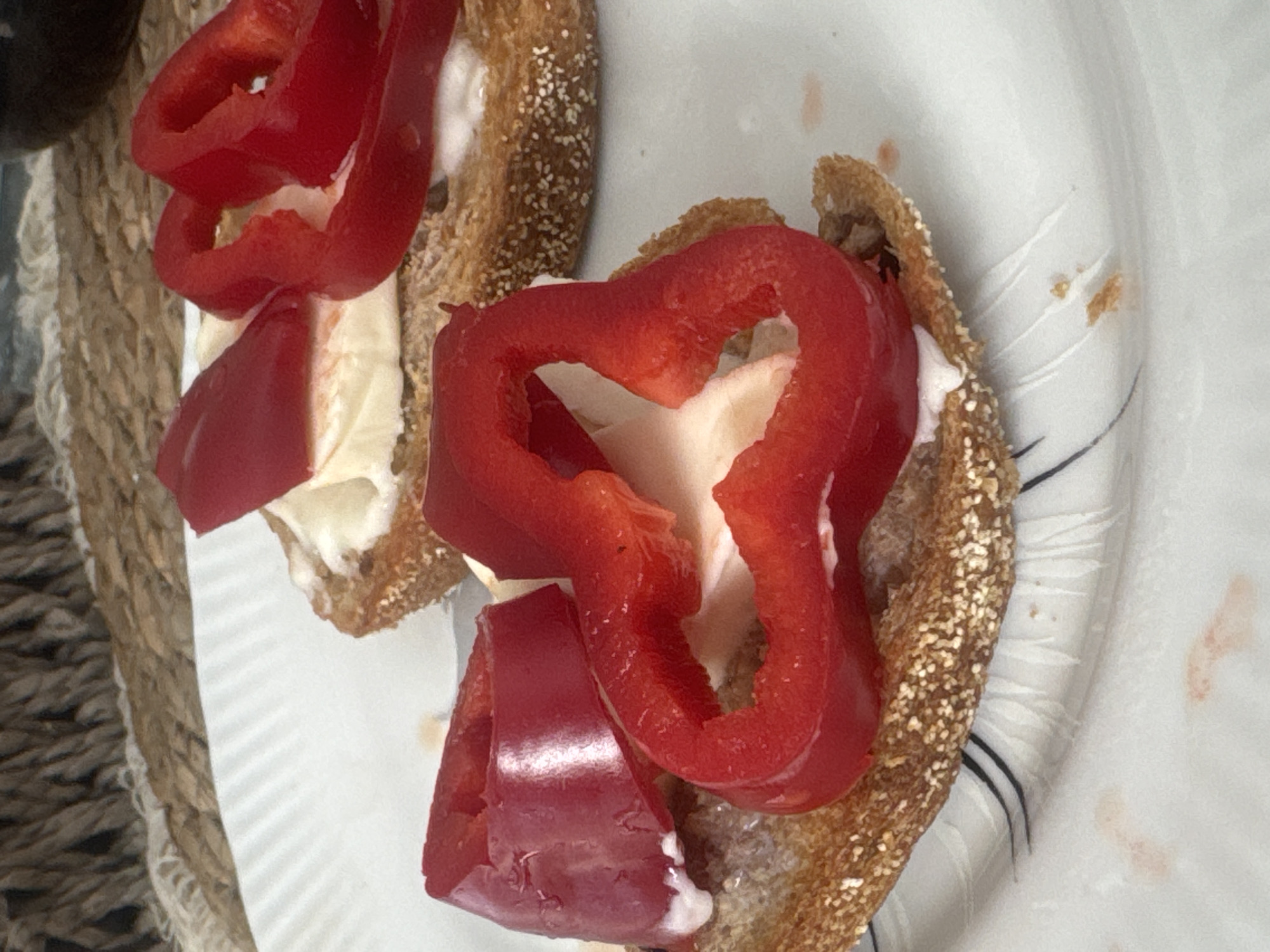 The image shows two pieces of toast topped with cream cheese and sliced red bell peppers, arranged on a white plate with black decorative lines. The plate is situated on a woven placemat. The food appears to be a light snack or appetizer. There are no people visible in the image. The background suggests an indoor setting, possibly a dining area. The image quality is clear, allowing for detailed observation of the food and plate. The location is Ramat Gan, Israel.