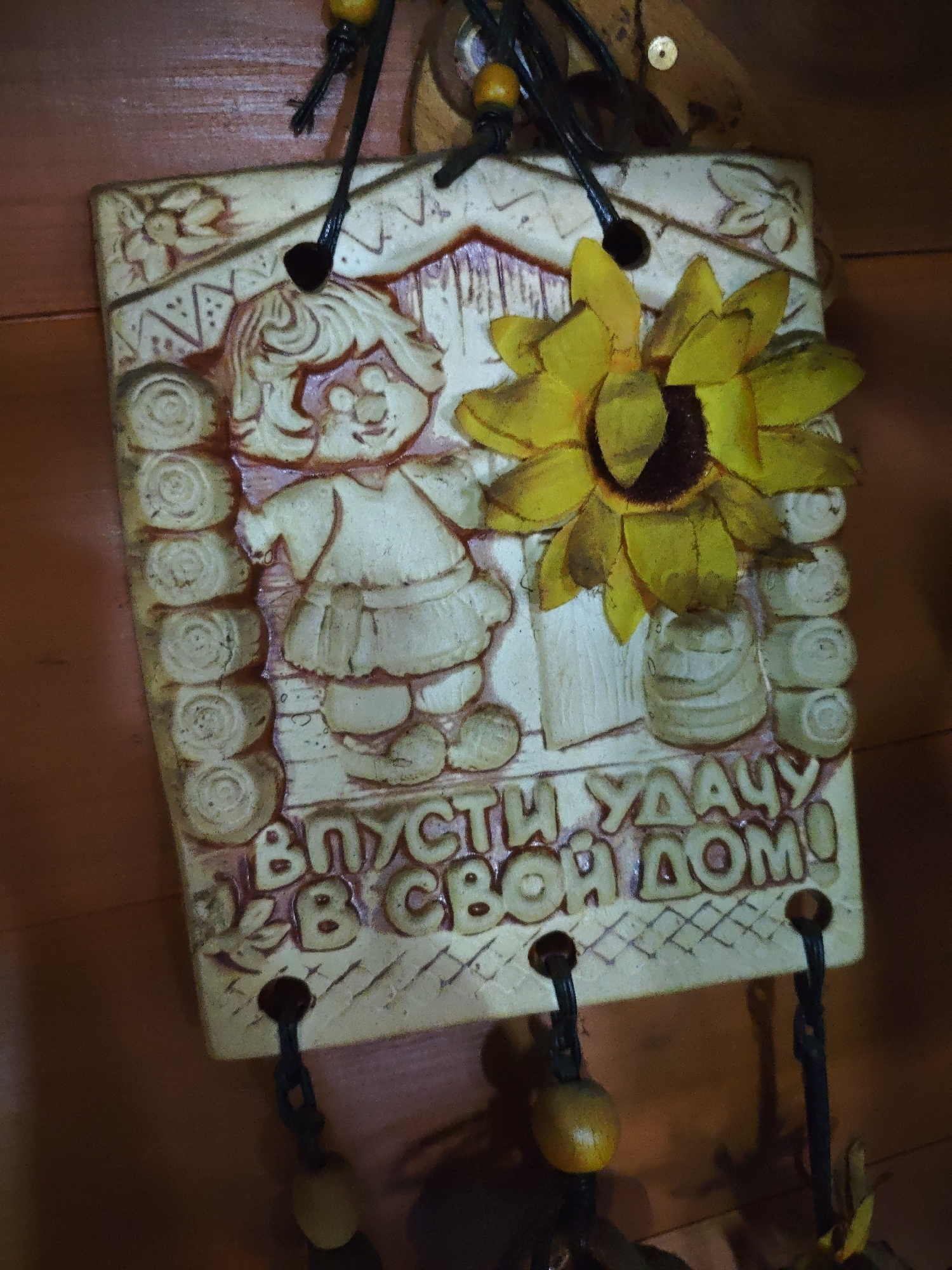 The image shows a close-up of a decorative wall hanging. It is made of light-colored material with embossed designs, including a cartoon-like girl, a sunflower, and what appears to be a bucket. Cyrillic text is present at the bottom, which translates to "Let luck into your home!"
The hanging has a string or rope with beads attached to the top and bottom for mounting. The background appears to be a wooden surface.
The location Боровково, Russia is not apparent in the image.