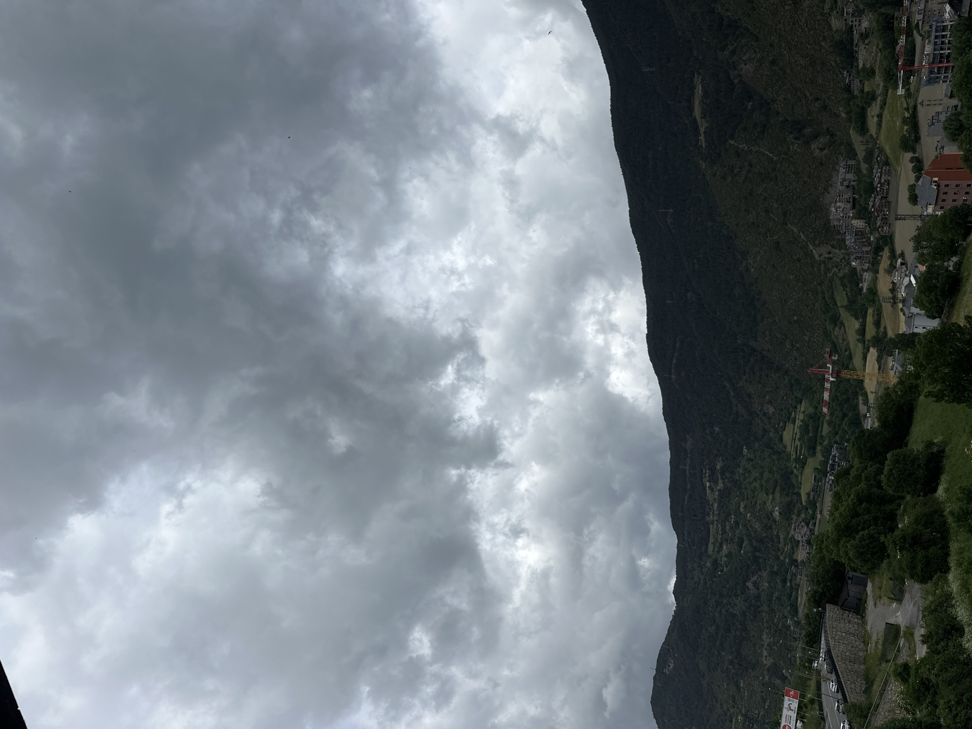 The image presents a panoramic view of a mountainous valley under a heavily overcast sky. The sky dominates the upper and left portions of the frame, filled with dense, dark grey clouds, suggesting either impending rain or a generally gloomy day. Small, dark specks, likely birds, are visible against the cloudy backdrop in the upper left.
A massive, dark green mountain rises steeply on the right side of the frame, its rugged slopes covered in abundant vegetation. Its peak extends beyond the frame or into the low-hanging clouds. Nestled in the valley below and extending across the middle and lower right of the image is a town or settlement. Various buildings, differing in size and style, are visible, interspersed with green patches of grass and numerous trees.
Roads wind through the valley, and several vehicles, primarily cars, can be seen on them, indicating active transportation. Notable man-made objects include at least two prominent yellow and red tower cranes, suggesting ongoing construction or development within the town. A stone retaining wall is visible along a roadside in the lower left. On the far bottom left, a large billboard with partially visible, illegible text and graphics can be discerned; "SANT" might be part of the text.
The scene depicts daytime under cloudy weather conditions. The overall impression is one of a human settlement integrated into a dramatic natural landscape, characteristic of a mountainous region like Encamp, Andorra. No specific human activity is clearly visible, but the presence of vehicles and construction implies a bustling, developed area.
