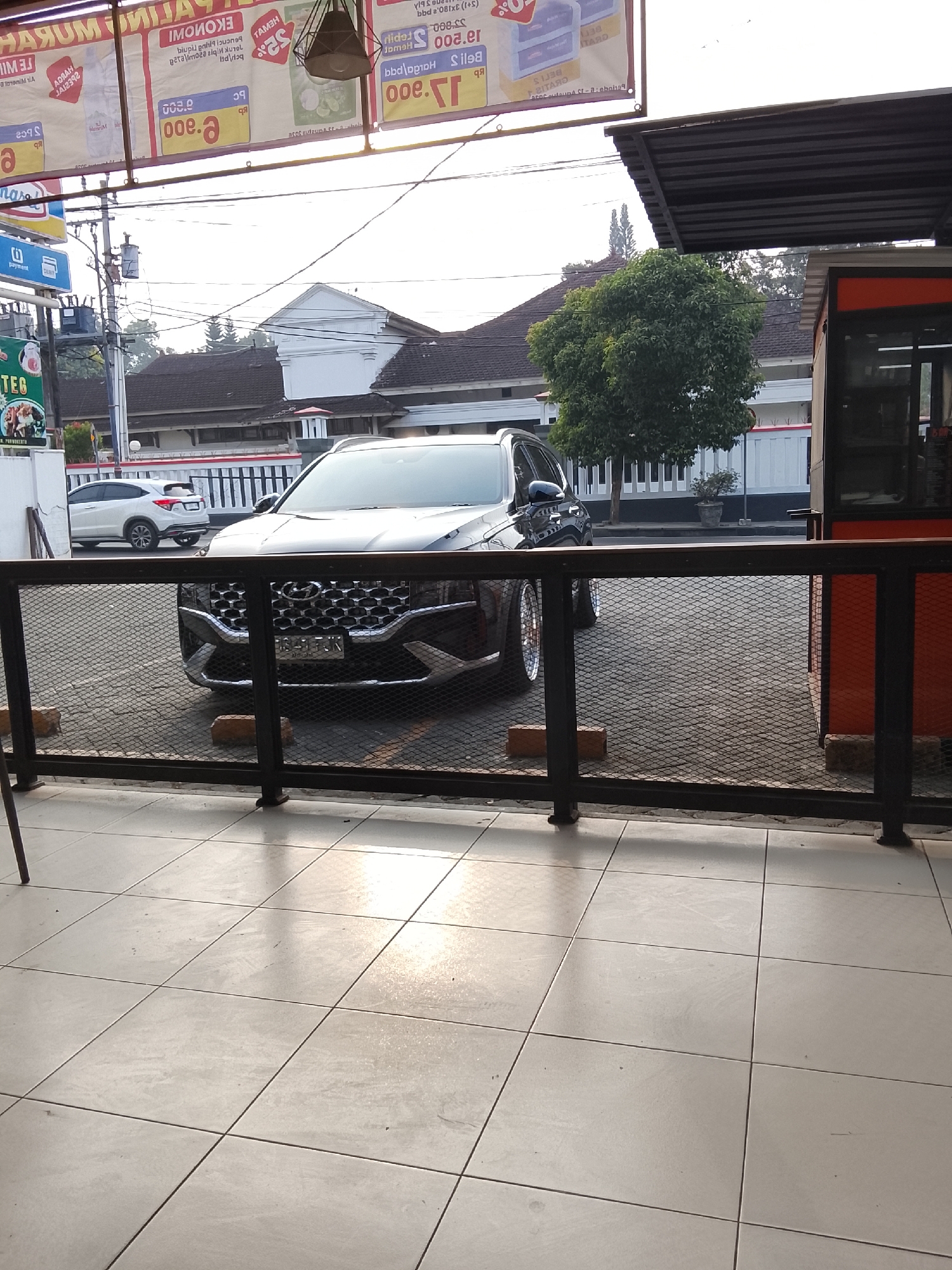 The image captures a frontal view of a dark-colored SUV parked on a paved lot in Kedungwuluh, Indonesia. The vehicle's distinctive grille and headlights are prominently featured. Behind the SUV, a white building with a white fence and a lush green tree are visible. Above the parking lot, banners with text and logos are suspended, hinting at a commercial area. A black metal fence runs across the foreground, partially obscuring the view of the car and its surroundings. The ground in front of the fence is tiled. The scene appears to be outdoors, with natural light illuminating the area.