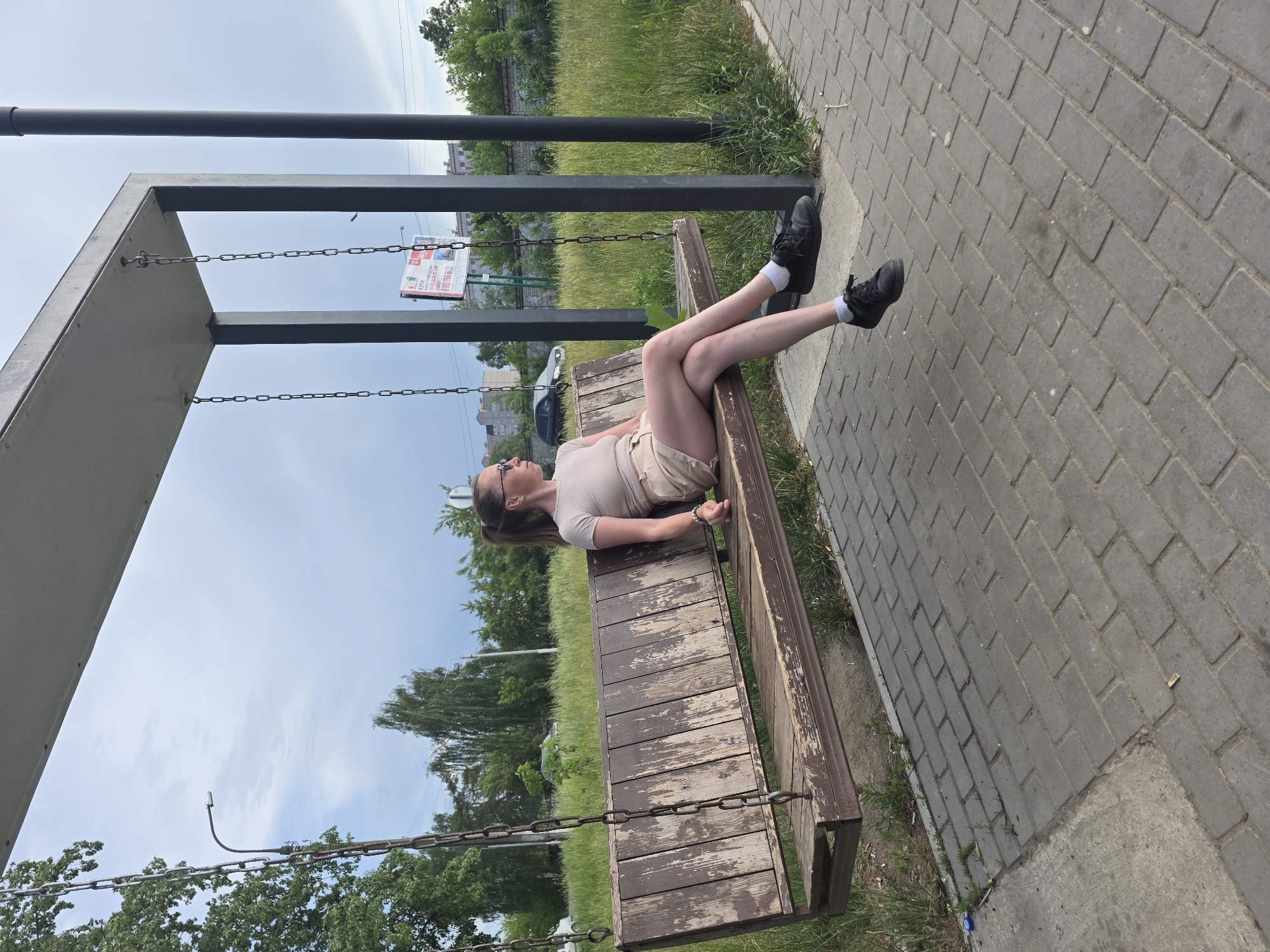 A full-length photograph shows a woman lying on her back on a swing bench in a park in what appears to be Chaikovsky, Russia. She wears a light tan t-shirt and shorts, white socks, and black shoes. Her hair is pulled back in a ponytail. The swing bench hangs from a black metal frame with chains. The ground is paved with gray bricks, and there is grass and trees in the background. A white sign with red and blue print and Cyrillic script is visible behind the swing.