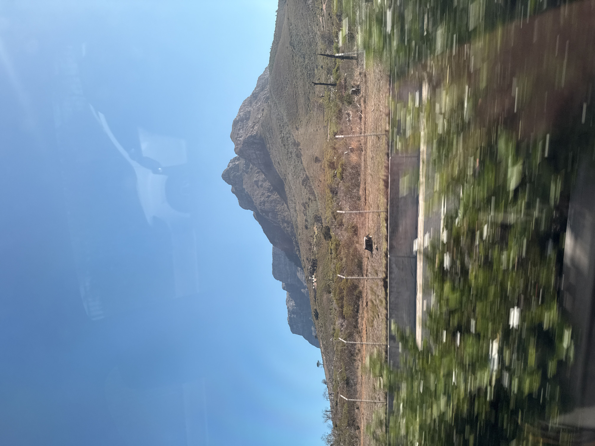 The image shows a mountain landscape taken from a moving vehicle, likely a car or train. A prominent, rocky mountain occupies the left side of the frame, set against a clear blue sky. The mountain appears to be composed of layered rock formations. At the base of the mountain, a patch of dry, brownish vegetation transitions into a fenced-off area alongside the road or railway. The fence consists of wooden posts and wire. On the right side of the image, blurry green foliage and reflections from the vehicle’s window obscure the background. Based on the landscape, it is likely that the mountain is located in the city of Cape Town, South Africa.