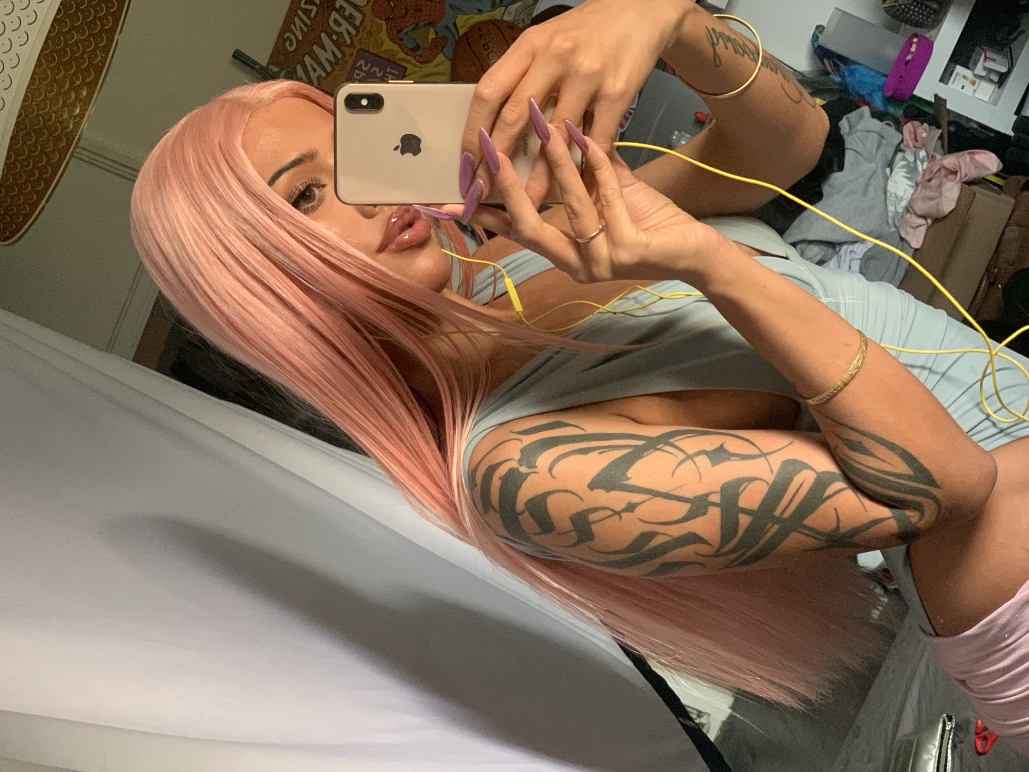 A light-skinned woman with a pink wig is taking a selfie using a silver iPhone. She is wearing a pale blue tank top. Her right arm has a large, intricate tattoo of stylized lettering. She is wearing gold bracelets on both wrists. She has long, purple nails. The background is cluttered and appears to be a bedroom. There is a messy desk and some clothing scattered around. The image does not contain any information that would allow for the location to be determined.