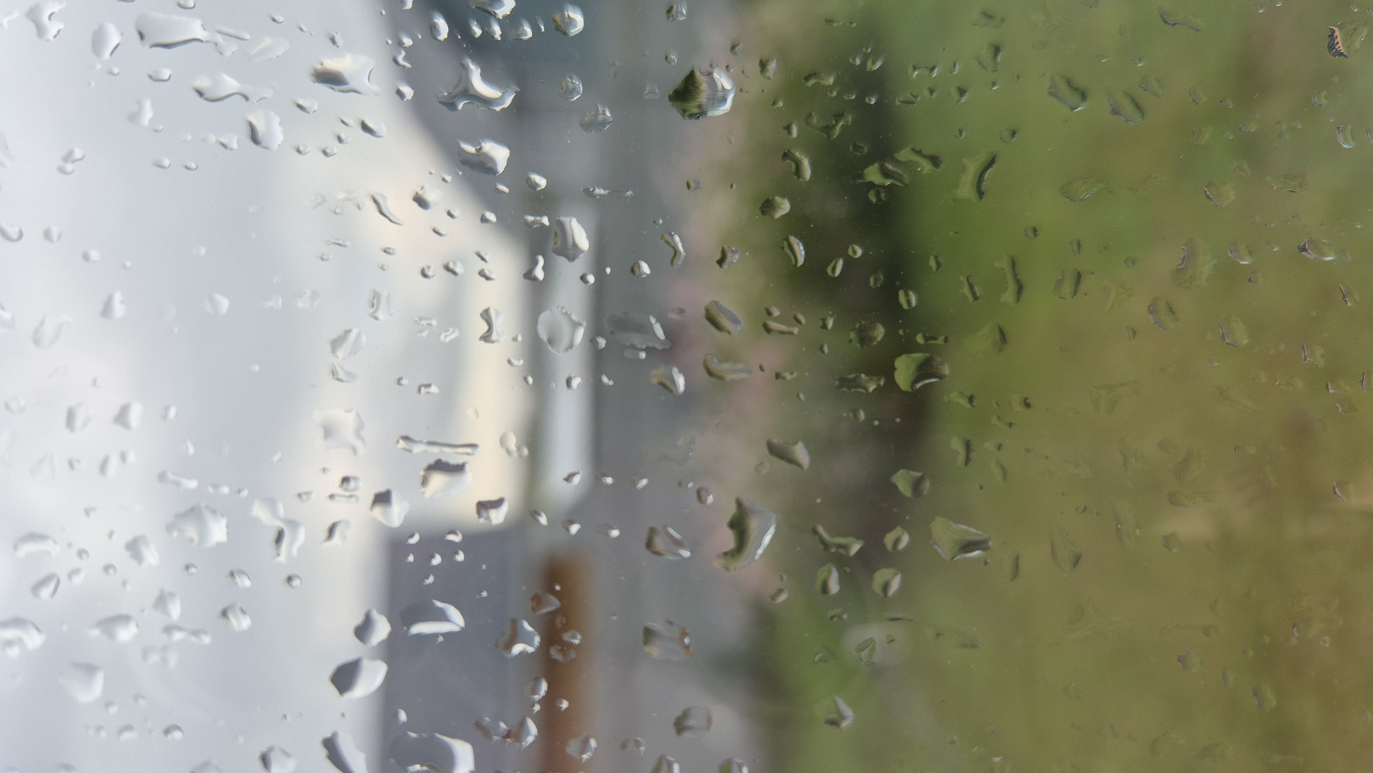 The image displays a close-up view of a window covered in water droplets, obscuring the scene outside. The blurred background suggests an outdoor environment with hints of structures, possibly buildings, and green foliage, indicating a suburban or rural setting. The overcast sky, characterized by diffused light, contributes to the muted color palette. The presence of water droplets on the window suggests recent rainfall or high humidity. The location is identified as Pomryaskino, Russia. No people or specific actions are visible due to the obstruction.