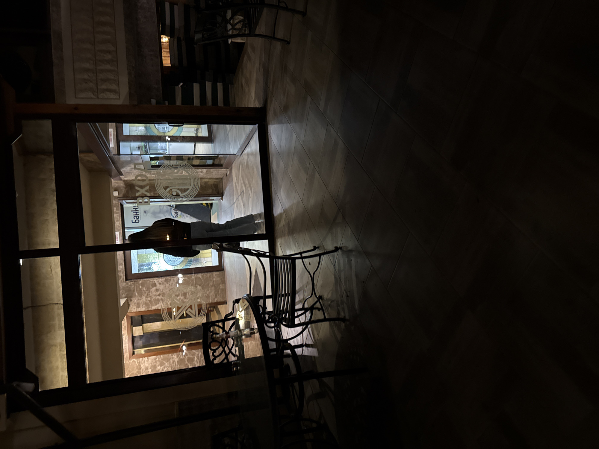 The image depicts a nighttime scene, likely an entrance to a business in Stara Zagora, Bulgaria. The viewer looks through a glass-paned door into an interior space. Reflected in the glass is a partial view of a person wearing dark clothing. The interior appears to be a restaurant or cafe with ornate decorations on the walls. To the right of the door, metal chairs are visible on a dark, wooden floor. The exterior lighting is dim. The door frame appears to be dark wood or metal. Text in Cyrillic script is visible on the glass of the door.
