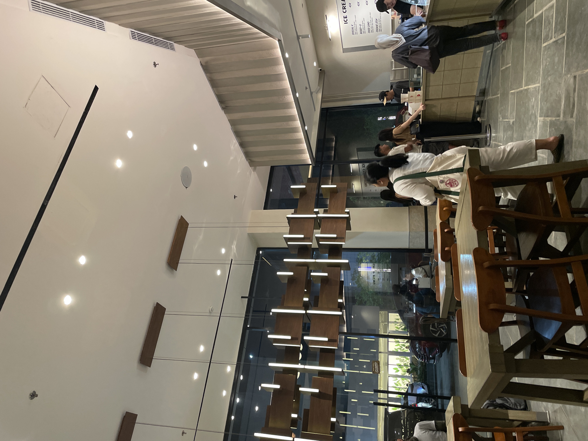 The image depicts the interior of an ice cream shop in North Jakarta, Indonesia. The shop has a high ceiling with recessed lighting and decorative wooden panels. A large, multi-tiered light fixture hangs down the center of the space, framed by large windows that offer a view of the outside.
Customers are seen ordering at the counter, and a barista is preparing orders. Tables and chairs are arranged for seating, and other patrons are present. The scene suggests an active and functional commercial ice cream operation.