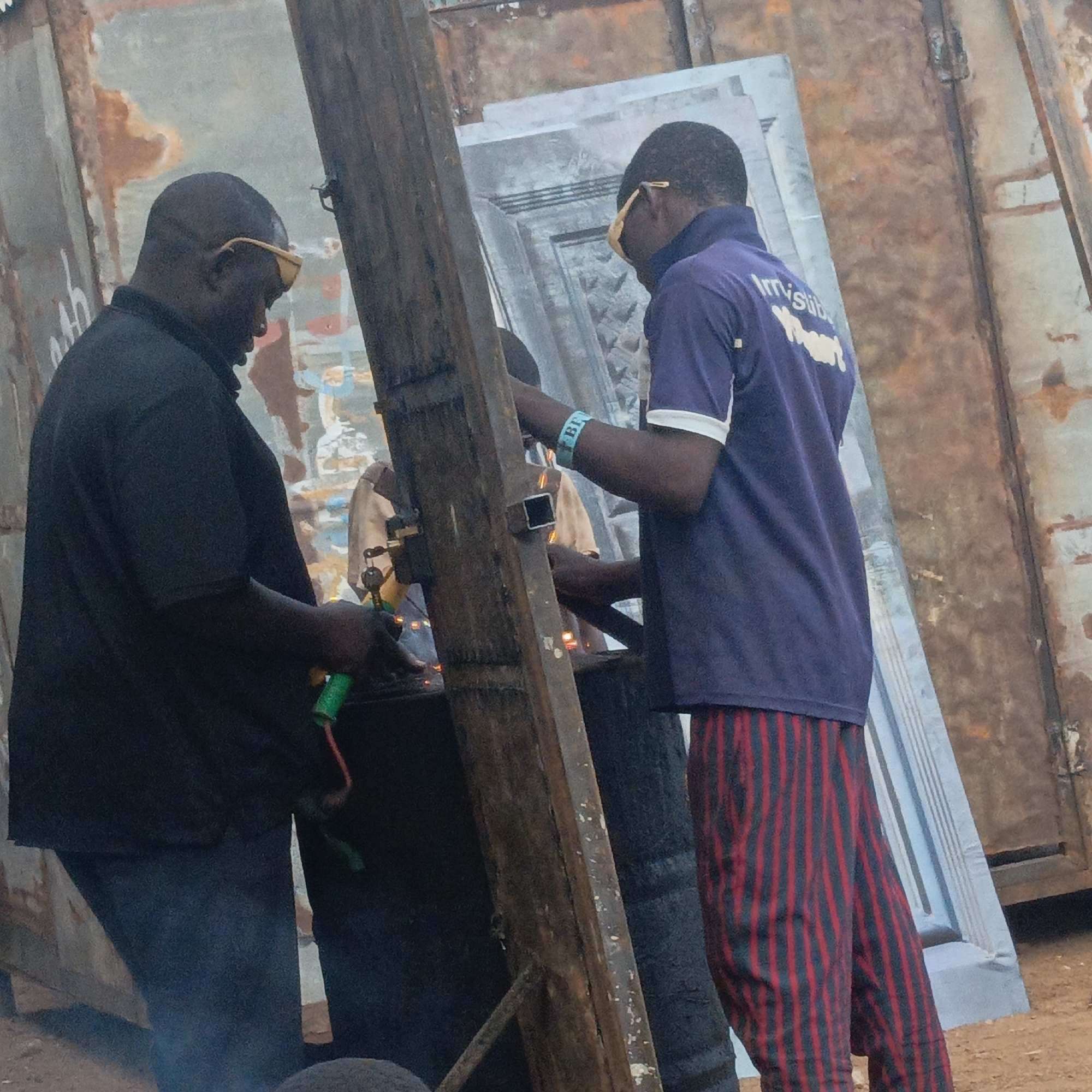 In Akwanga, Nigeria, two men are welding metal. Both men are wearing safety glasses. One man, in a black shirt and jeans, is operating a welding tool with a green handle. The other man, wearing a purple shirt with white lettering and striped pants, is holding a metal bar. A piece of metal is mounted on a wooden post that serves as a makeshift workbench. Welding sparks are visible, suggesting active welding. In the background, there are rusted metal sheets or containers, indicating an industrial or construction site. A third individual in a light-colored shirt can be seen standing behind the metal being welded.