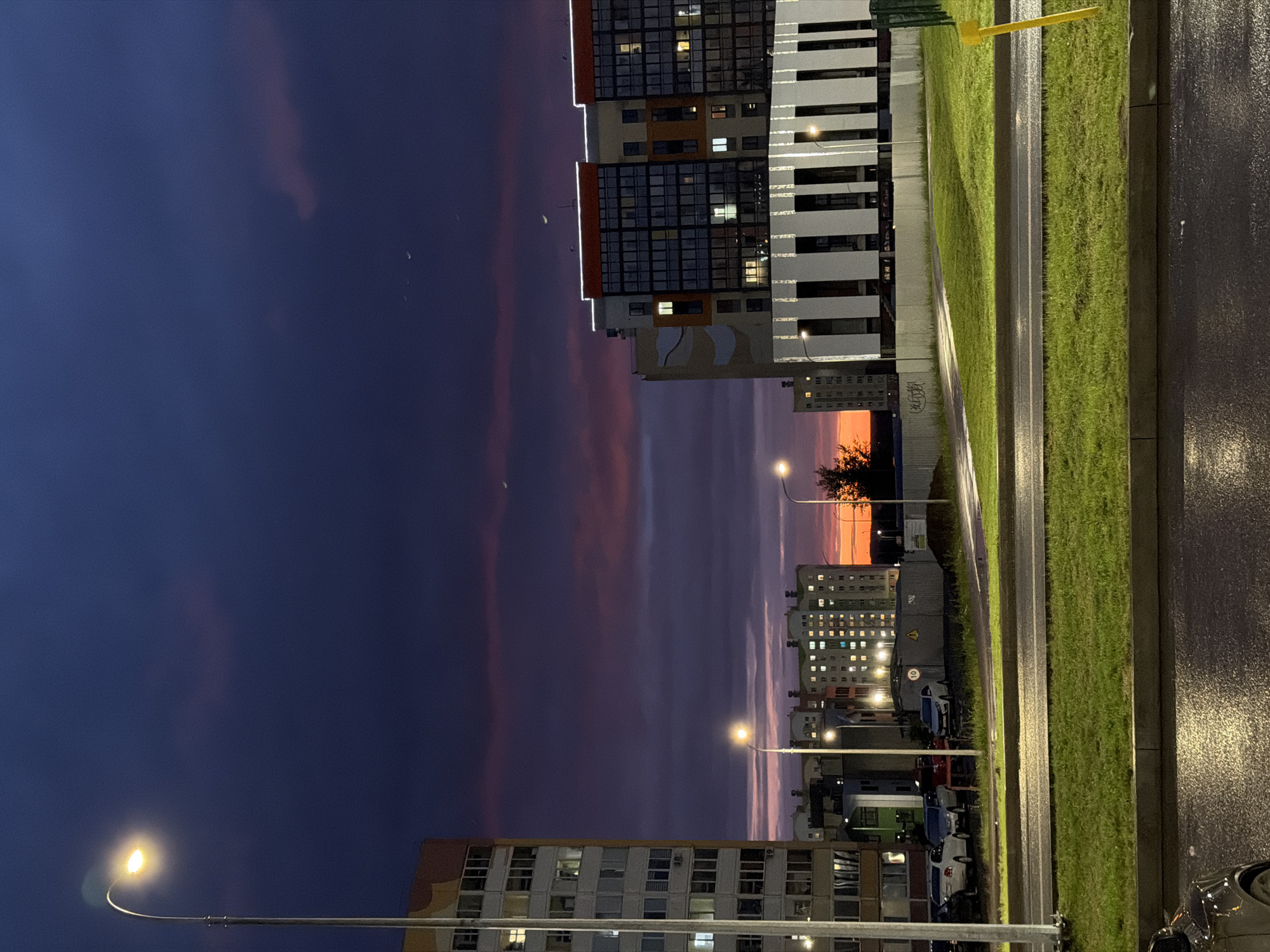 The image depicts a street scene in Nizhnekamsk, Russia, at dusk. The sky is a deep purple, transitioning to lighter shades of pink and orange near the horizon, indicating the sunset. Scattered clouds are visible, tinged with the colors of the setting sun.
Dominating the right side of the frame are modern apartment buildings with numerous lit windows, suggesting that people are home and settled in for the evening. The architecture appears to be contemporary, with balconies and varied facade designs. One building has a striking white and black striped pattern. A well-maintained patch of green grass runs alongside the buildings, separated by a paved area that appears to be a sidewalk or road. The ground is wet, hinting at recent rain or a damp atmosphere.
Streetlights illuminate the scene, casting a warm glow on the wet pavement and the surrounding grass. A yellow pole, possibly a part of a street sign or utility pole, stands out against the darker sky. In the distance, more apartment buildings are visible, also dotted with lights. The overall mood is serene and peaceful, capturing the quiet transition from day to night in a residential urban environment. No people are prominently visible, focusing the attention on the architecture and the natural beauty of the sunset.