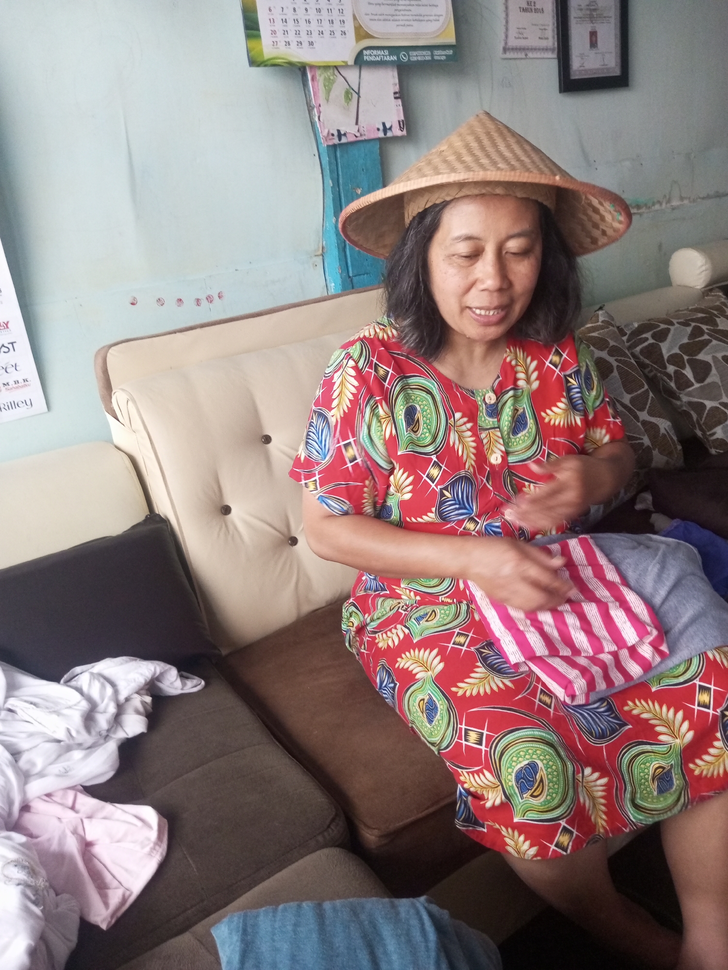 The image shows a woman wearing a conical hat and a colorful red dress with a floral pattern, sitting on a couch in what appears to be a living room in Andongsili, Indonesia. She is holding a pink and white striped cloth in her hands and looking down at it. The couch is upholstered in beige and brown fabric, and there are several cushions and some folded clothes on it. The wall behind the couch is painted light blue and has a calendar and some framed pictures hanging on it. The overall impression is one of a casual, domestic setting.