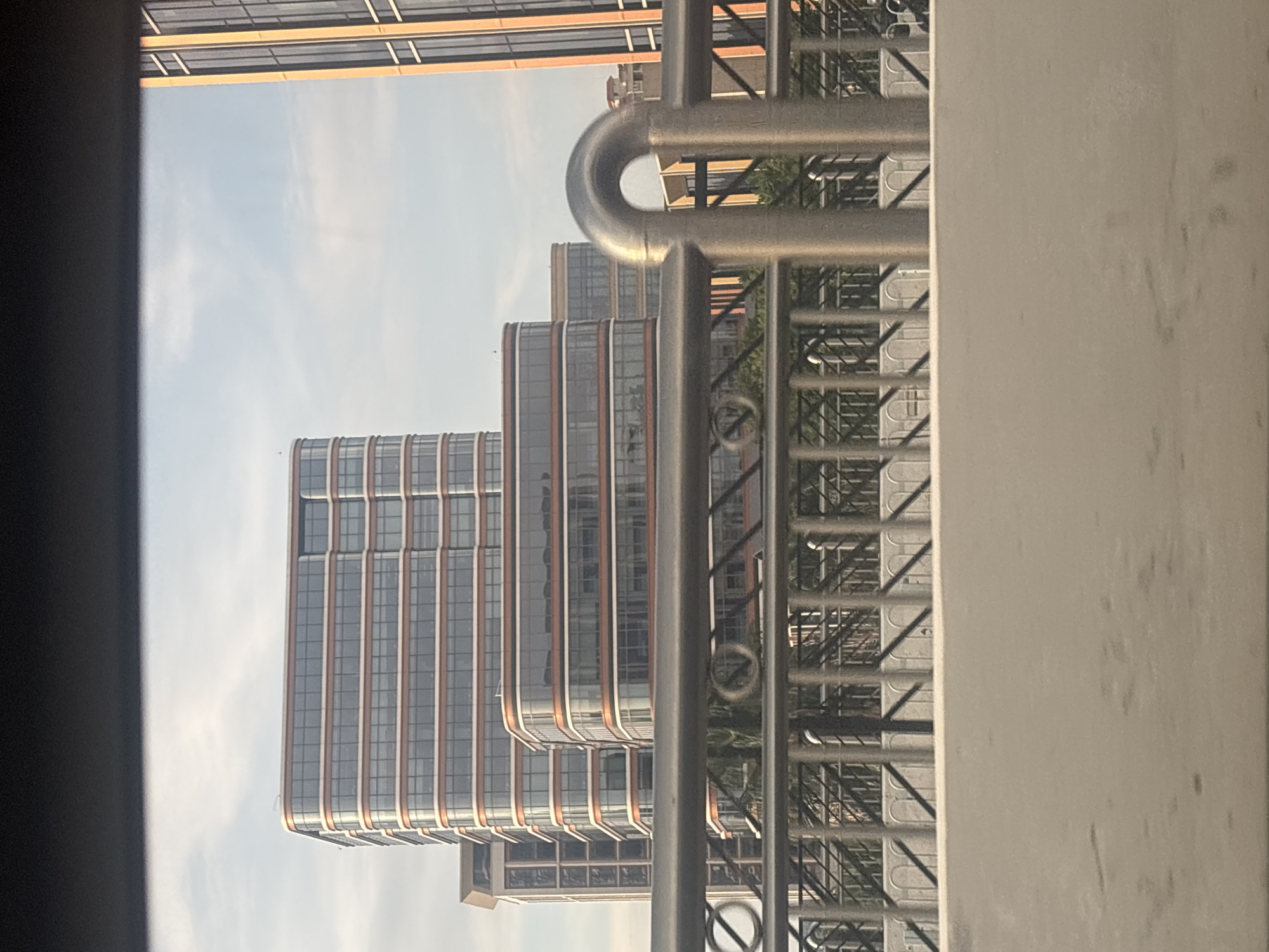 The image shows a view of modern buildings in Baiyun District, China, from an elevated position, possibly a bridge or overpass. The foreground is dominated by a light-colored concrete barrier. Behind this barrier is a metal railing with vertical posts and horizontal bars, some of which have decorative, curved elements. Beyond the railing, several multi-story buildings are visible. One prominent building on the left is tall and features a facade with horizontal bands of glass windows and what appear to be bronze or copper-colored trim. To its right, a shorter, curved building with a similar facade design is situated. Further to the right, another tall building with a similar facade is partially visible. The sky above is light blue with scattered white clouds. The overall scene depicts an urban landscape with contemporary architecture.