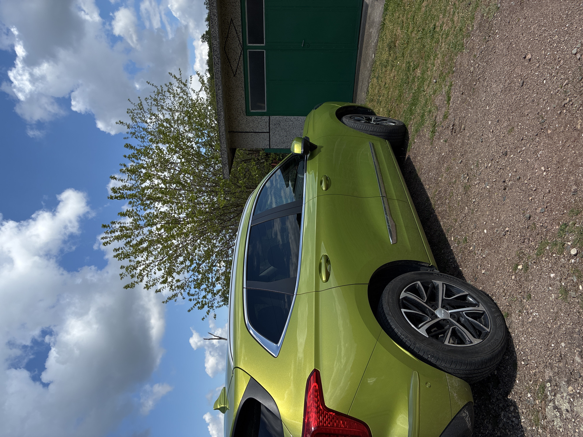 The image shows a light green car parked on a gravel surface near a small, single-story building with green doors. The building appears to be a garage or storage shed. A tree with budding green leaves stands near the car. The sky is blue with scattered white clouds. The location is implied to be Bokilovtsi, Bulgaria. There are no people visible in the image. The primary subject is the car, its color, and proximity to the building. The environment suggests a rural or suburban setting.
