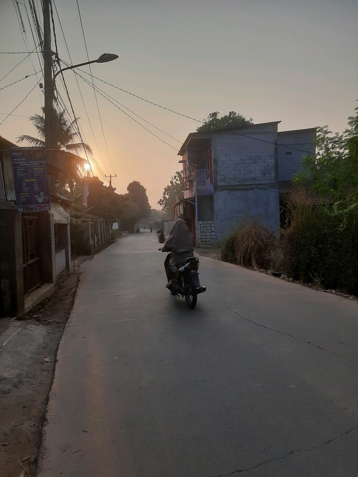 In Gandoang, Indonesia, a person rides a motorcycle down a street during sunrise. The road is paved and has visible cracks. On the left side of the road, there is a sidewalk and a building with a sign that reads "MINI ATM TRANSFER - TARIK TUNAI - PEMBAYARAN". There are utility poles and many wires above the road. On the right side of the road, there is a two-story building constructed with grey cinder blocks, surrounded by lush green trees and tall grasses. The sun is low on the horizon, casting a warm glow on the scene.