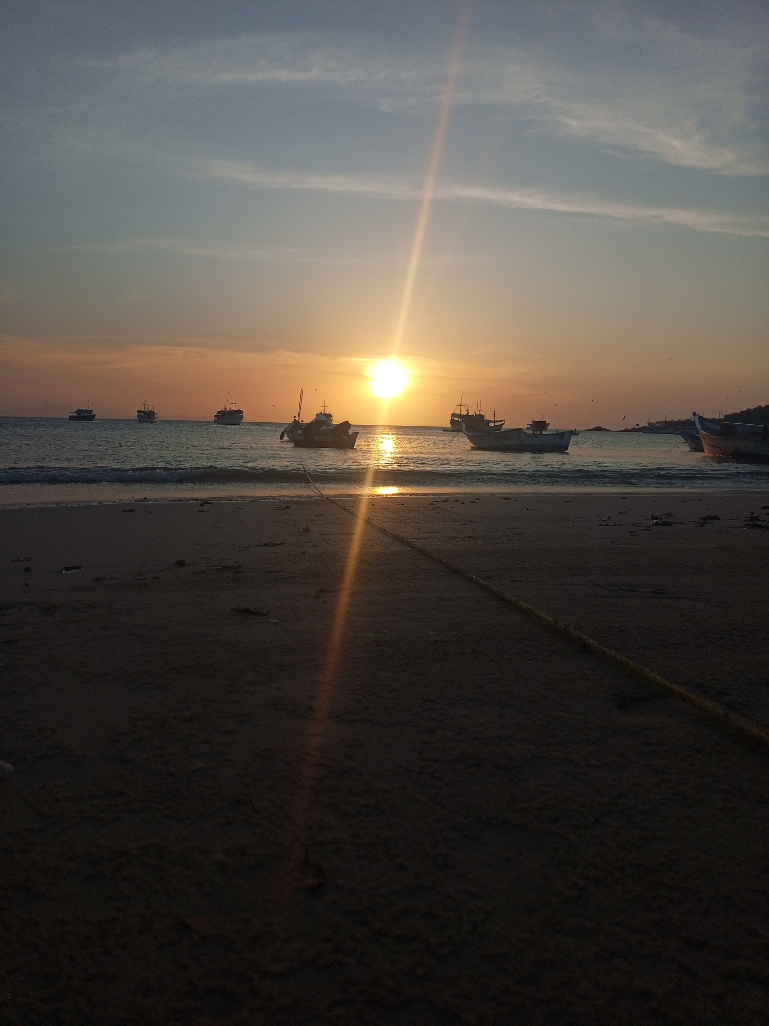 The image depicts a sunset scene at a beach in Juan Griego, Venezuela. The sun is setting over the ocean, casting a bright glare and a golden light across the water and sand. Several fishing boats are anchored in the calm water near the shore. The beach sand in the foreground is dark, with a rope stretched across it. The sky transitions from blue to orange near the horizon. Some birds are flying in the distance.