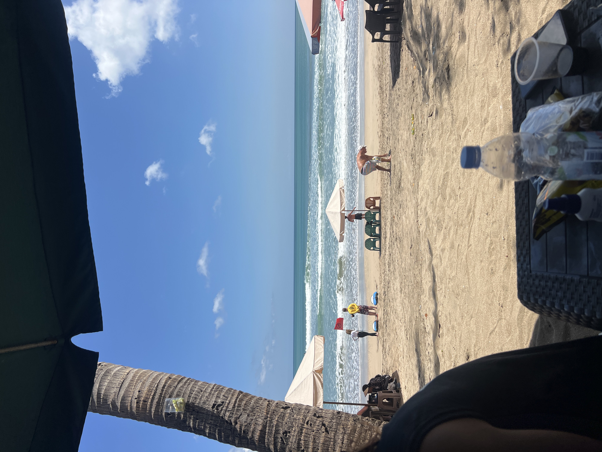 The image captures a sunny day at Kuta Beach, Bali, Indonesia. In the foreground, a dark umbrella partially obscures the view, with the textured trunk of a palm tree to its left. A table is visible in the lower right corner, holding a plastic water bottle, a plastic bag, a phone, and a cup. The beach itself is sandy, with several beach umbrellas and chairs scattered across it. In the middle ground, a man in patterned shorts bends down on the sand, facing away from the camera, while another person in a yellow shirt and a person in a white shirt stand near the water's edge, holding a red flag. Two blue flotation devices are on the sand near them. Further back, the turquoise ocean meets the horizon under a bright blue sky dotted with white clouds. The waves are breaking on the shore, creating a white foam.