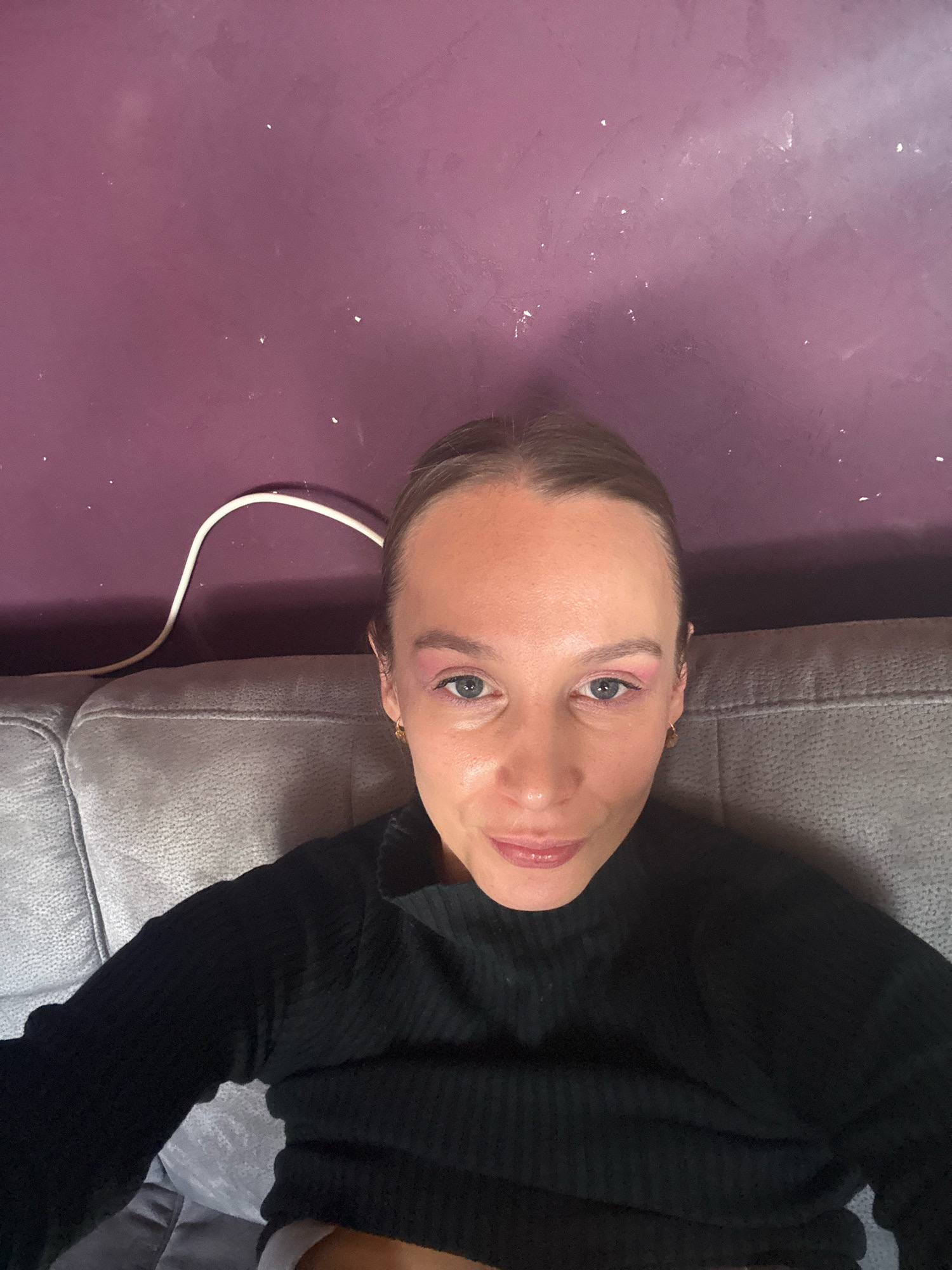 In a dimly lit room, a woman with light brown hair pulled back sits on a gray sofa. She is wearing a black long-sleeved shirt. Her makeup includes pink eyeshadow and lip gloss. Behind her, a purple wall with a white cable draped across it is visible. The scene appears to be within a residential setting, possibly a living room, in Überherrn, Germany.