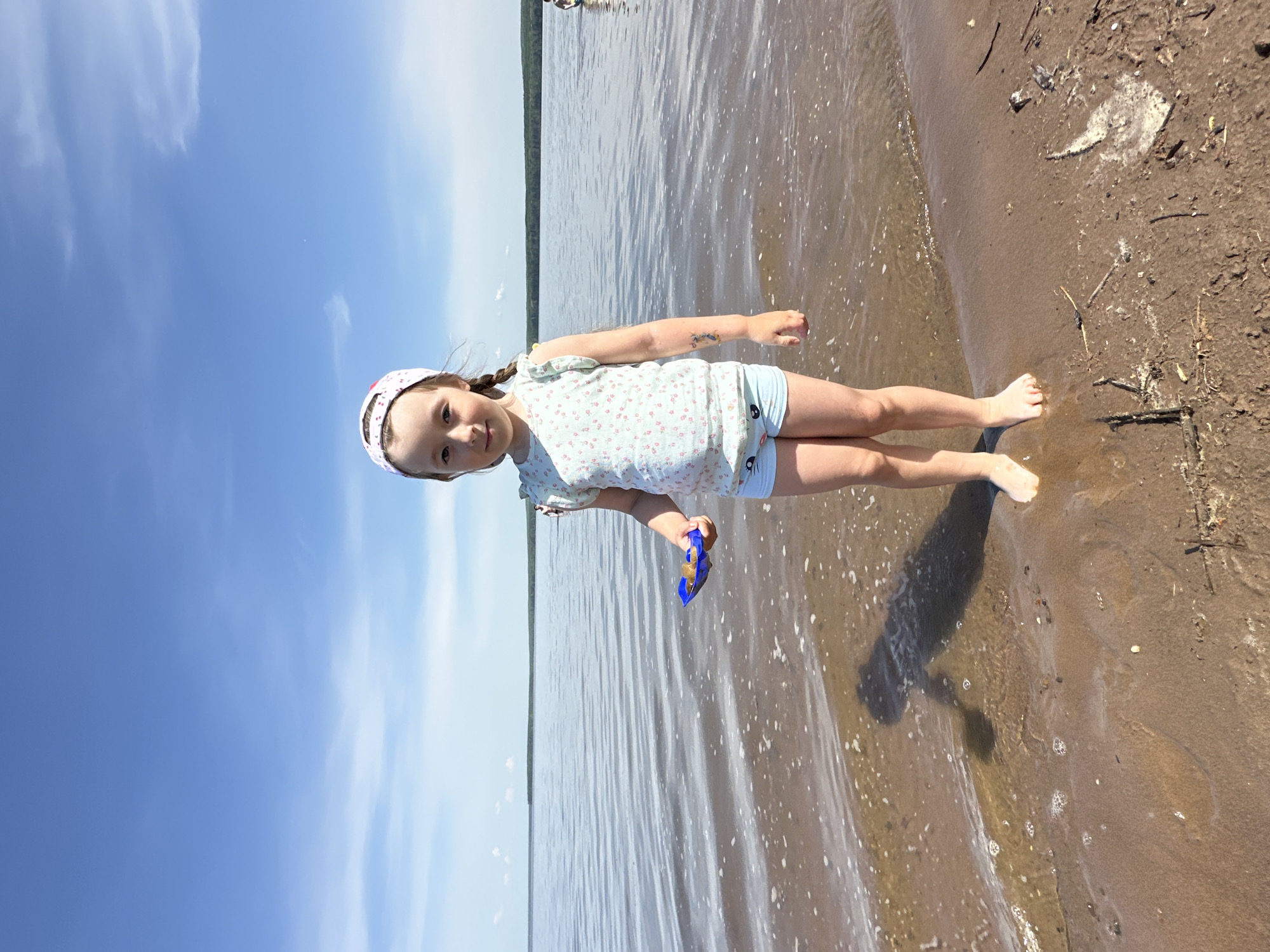 A young girl stands on the shore of a body of water, possibly the Kama Reservoir, in Chaikovsky, Russia. She wears a light-colored, floral-patterned shirt, blue shorts, and a patterned headband. She holds a blue plastic toy in her right hand. The water laps at the shore, and the sky is partly cloudy. To the right of the girl, there is sand with some debris. In the background, across the water, there is a line of trees.
