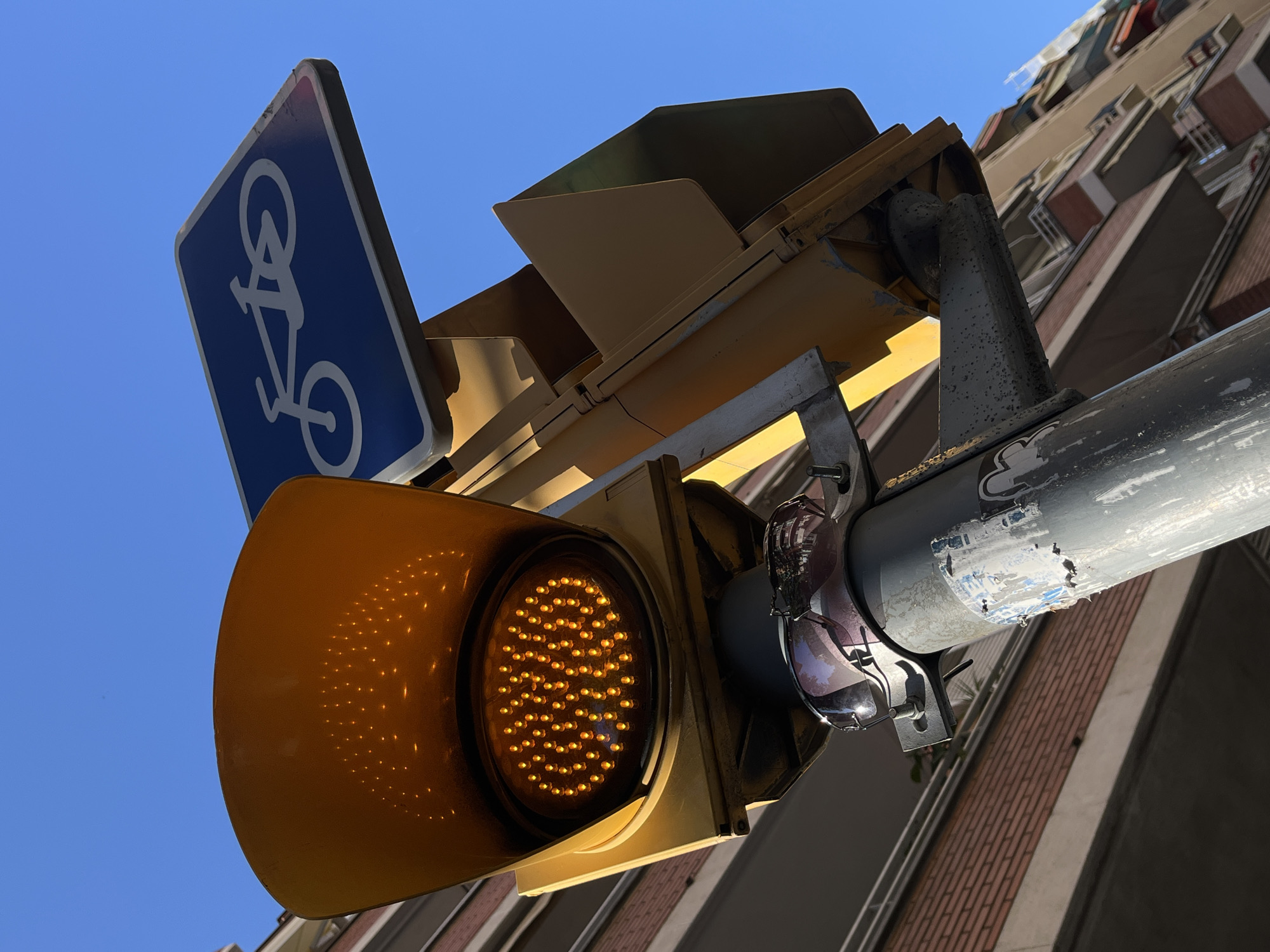 A low-angle shot depicts a yellow traffic light illuminating against a clear blue sky, with a bicycle route sign positioned above it. A building with a beige and brown facade is visible in the background. Sunglasses hang off of the supporting arm of the light.