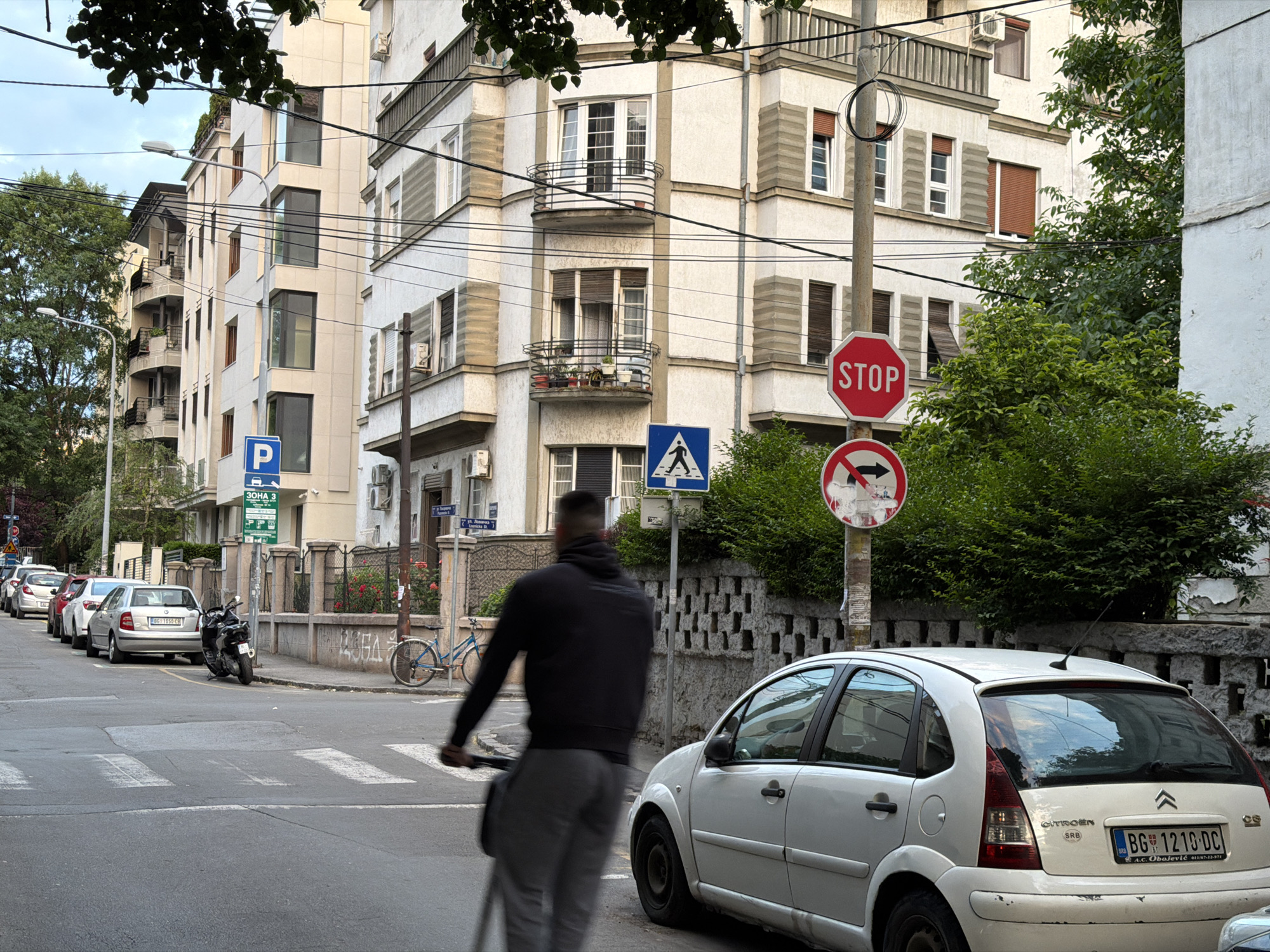 The image captures a street scene in the Vracar Urban Municipality, Serbia. The backdrop consists of multi-story buildings with architectural details like balconies and window shutters.
A man on a scooter is crossing a crosswalk. Cars are parked along the left side of the street, and a bicycle leans against a fence.
Several street signs are visible, including a parking sign ("P" and "30HA 3"), a pedestrian crossing sign (the iconic symbol of a person walking across a crosswalk), a stop sign, and a 'do not enter' sign. A white Citroen car is parked on the right side of the street. Its license plate reads "BG 1210 DC."