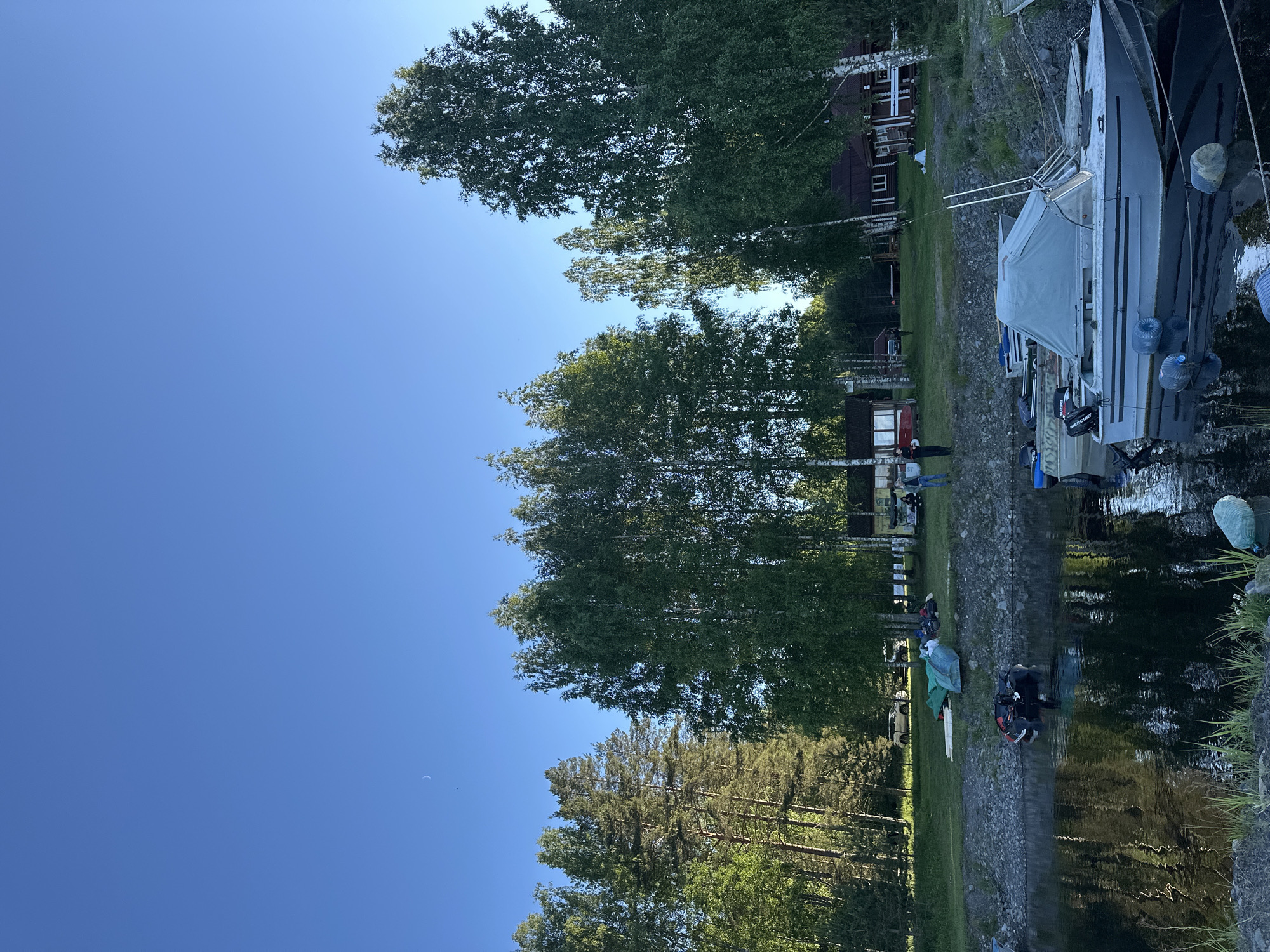 This is a photograph taken in Yanisyarvi, Russia, depicting a serene waterfront scene. The sky is a clear, bright blue with a faint crescent moon visible in the upper left quadrant. The foreground is dominated by a body of dark water reflecting the surrounding trees and boats. Several boats are docked along the shore. The closest boat is a light-colored motorboat with a grey cover and two vertical antennas. Further back, more boats, some with outboard motors, are moored.
The shore is lined with a rocky embankment. Beyond the embankment, a grassy area slopes upwards, dotted with trees. A cluster of birch trees with white bark and green leaves stands prominently in the middle ground. Behind the trees, several rustic wooden buildings with red roofs are visible. To the left, a dense forest with tall pine trees borders the scene.
On the grassy area near the shore, a few people are gathered. Two individuals are standing near a building, and another person can be seen further in the distance. Various objects, including what appears to be green tarpaulins and a deflated blue raft, are spread on the ground. A white vehicle is parked on the grass in the background, partially obscured by trees. The overall impression is one of a tranquil summer day in a rural, lakeside environment.