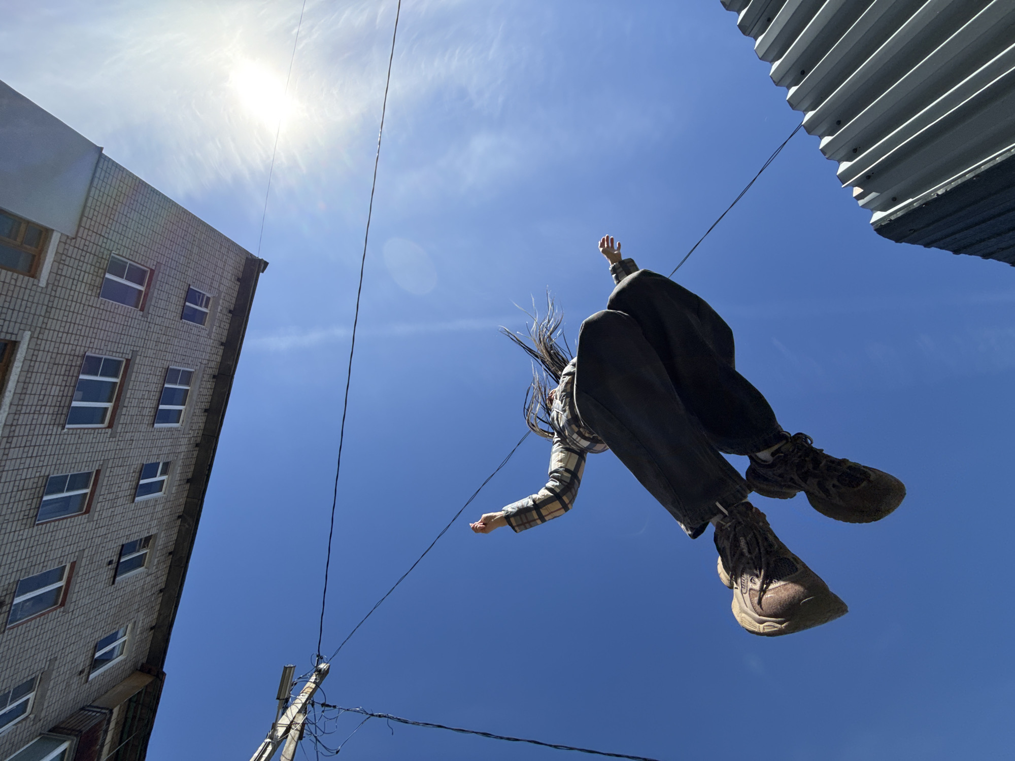 A low-angle outdoor photograph captures a person seemingly suspended in mid-air against a vibrant blue sky, framed by urban architecture. The bright sun, positioned in the upper left, casts a strong glare and produces visible rays through wispy clouds.
The central subject is a person, visible from the torso down, with their long dark hair appearing windswept. They are dressed in dark pants, a plaid long-sleeved shirt, and sturdy, dark-colored athletic shoes or boots. Their arms are outstretched, and their body is angled, contributing to the dynamic impression of being airborne, possibly jumping or falling.
On the left side of the frame, a multi-story building with a grey, brick-like exterior and numerous rectangular windows is angled upwards, indicating the low photographic perspective. On the upper right, a corrugated metal structure, possibly part of a roof or awning, cuts into the frame. Several thin black wires crisscross the sky, with one appearing to lead down towards a utility pole visible in the lower left portion of the image.
The scene takes place on a clear, sunny day, likely during mid-day given the intensity of the sunlight. The architecture, including the brick building and utility pole, suggests an urban environment, consistent with Rusanovo, Russia. The overall impression is one of dynamic motion and an unusual perspective, creating an artistic or playful representation of flight or suspension. No visible text is present in the image.