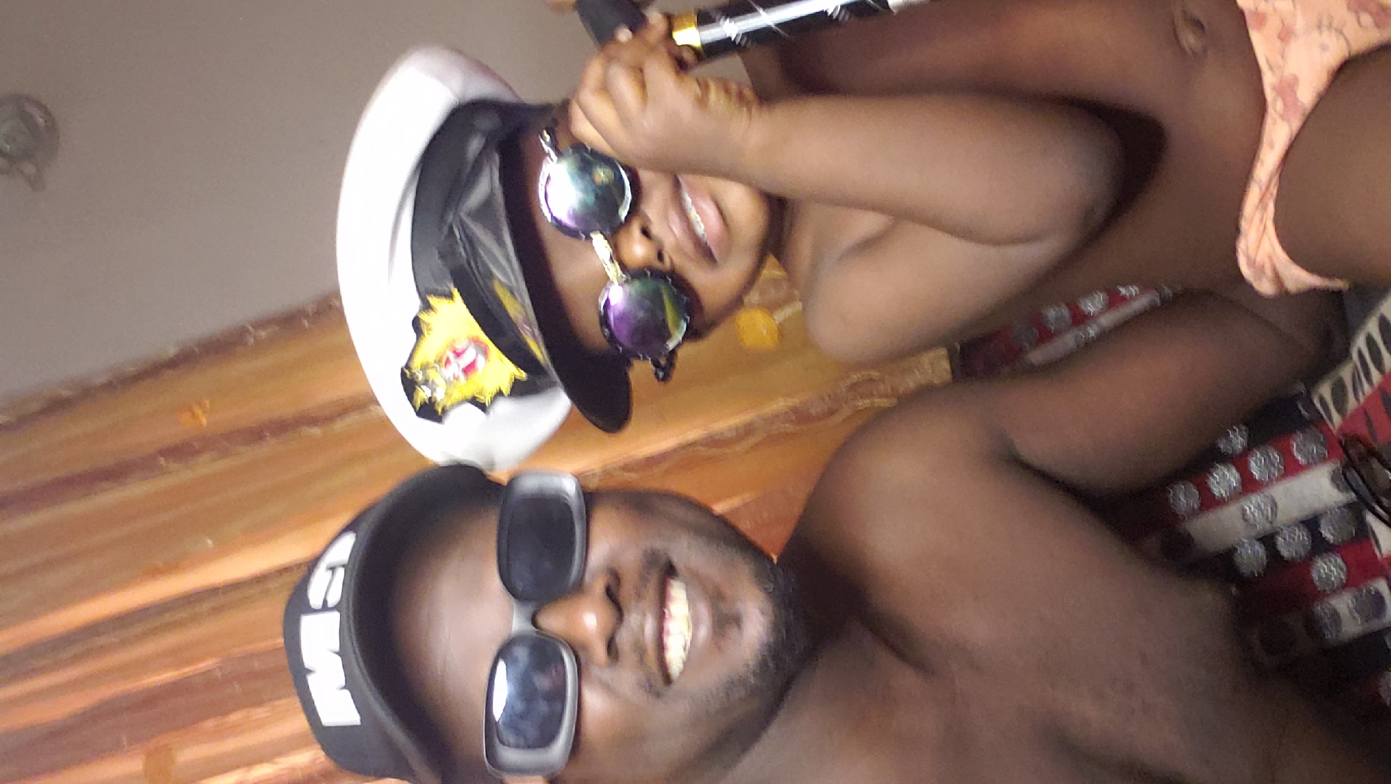 The image captures two individuals with dark skin tones in a close-up indoor setting, likely a selfie. Both subjects are smiling broadly, conveying a cheerful mood.
In the foreground, a man with a beard is seen from the chest up. He wears dark, rectangular sunglasses and a black baseball cap featuring white stylized text, possibly "GvM" or "GeM". His upper body is bare. Partially visible beneath him is a cushion or pillow with a distinct red, white, and black pattern.
Behind him, a younger individual, likely a child, is visible from the waist up. They are wearing round, reflective sunglasses and a white and black captain's hat adorned with a yellow and red emblem. This person is holding a dark object, possibly a bottle or tube with a metallic top. Their upper body is also bare, with light-colored undergarments visible around their waist.
The background shows a wooden or wood-patterned surface and a light-colored wall or ceiling, where a faint circular fixture can be seen. The playful attire and happy expressions suggest a casual, relaxed, and joyful moment, possibly a photo taken while lounging. The scene takes place in Sabon Tasha, Nigeria.