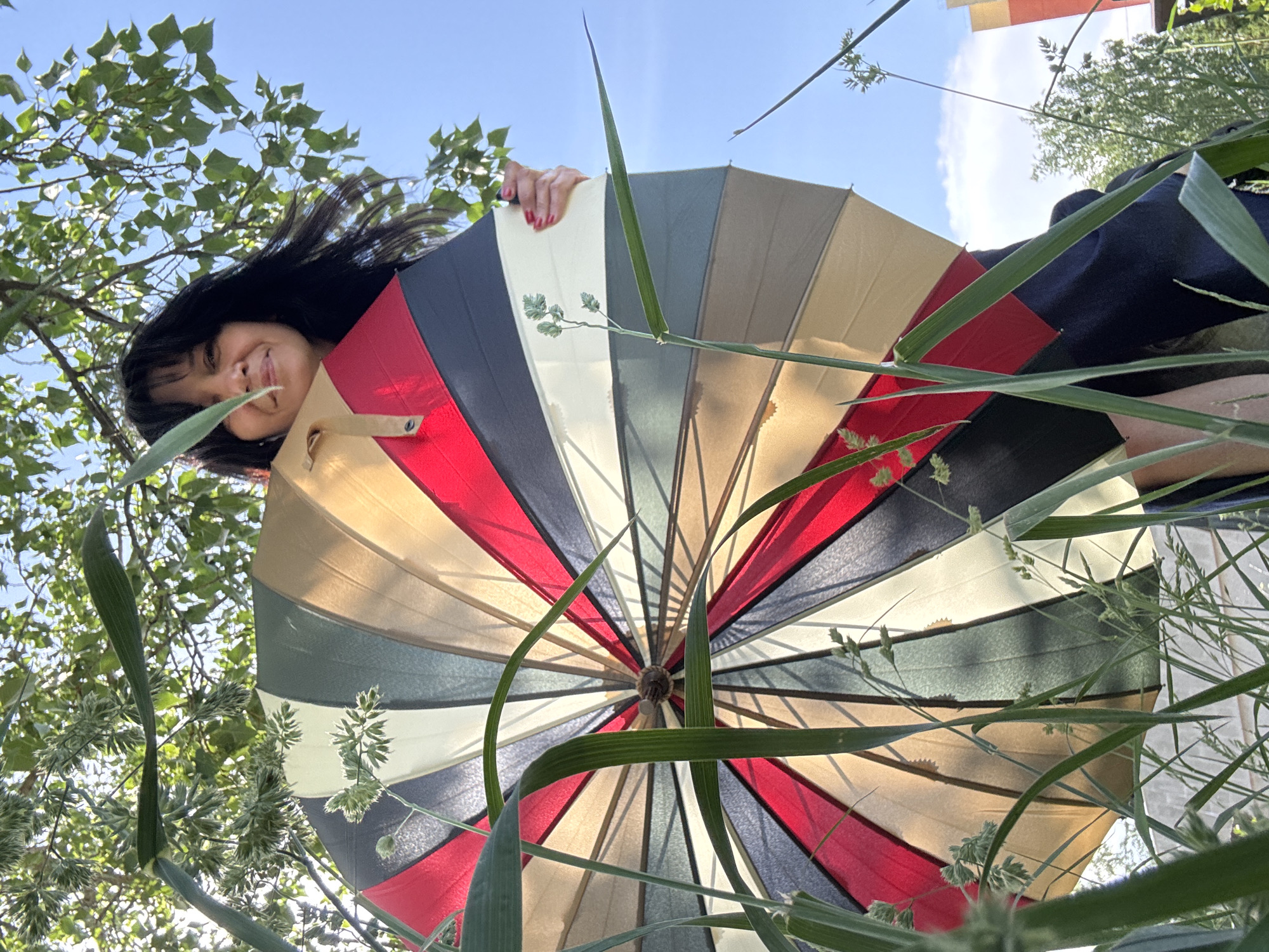 A low-angle shot depicts a woman partially obscured behind a multi-colored umbrella, likely outdoors in Chaikovsky, Russia. The woman has dark hair and is smiling slightly. The umbrella is composed of various colored panels including red, black, white, beige, and green. Tall grass and foliage surround the umbrella. Above the foliage is a blue sky.