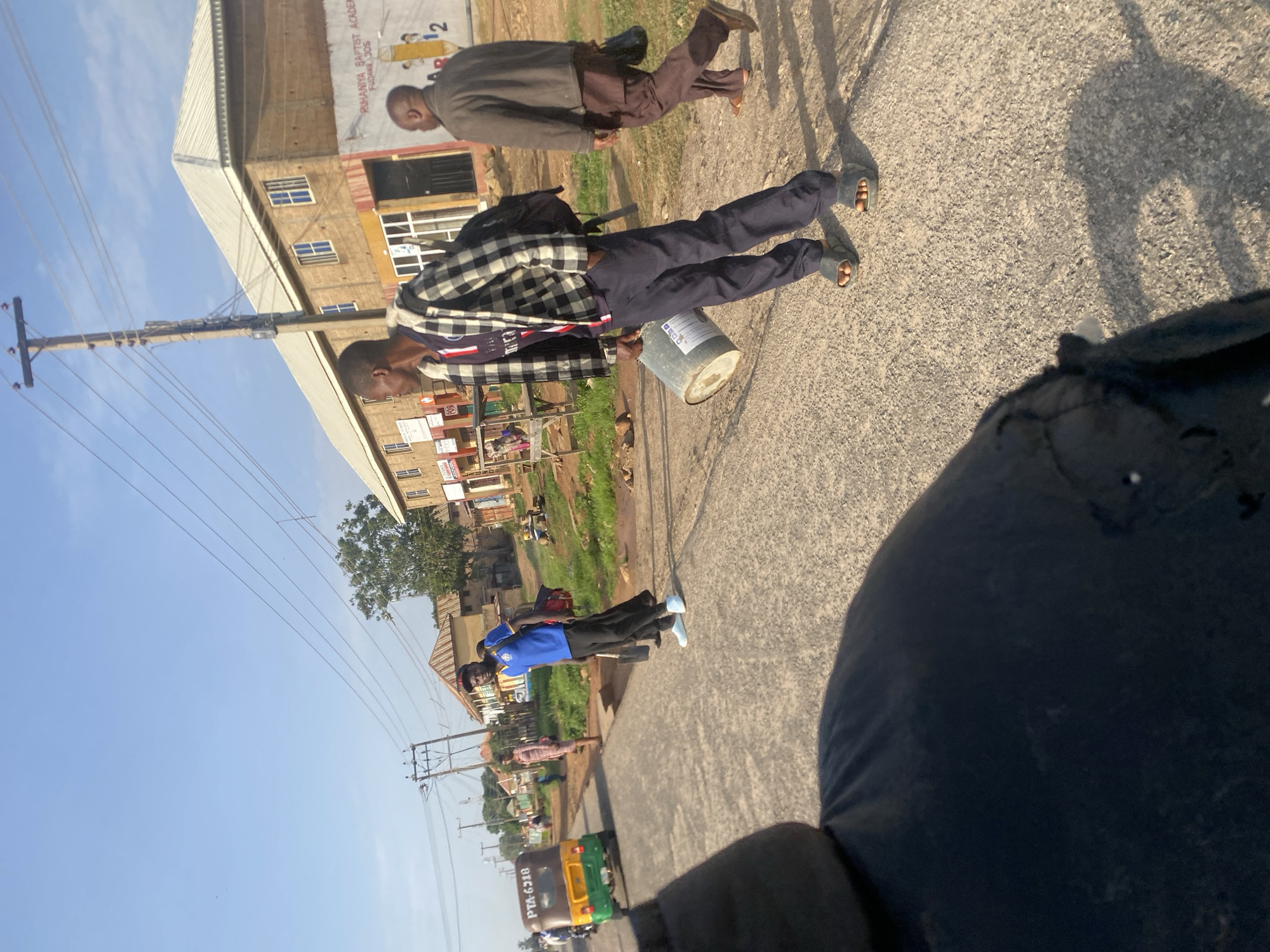 The image depicts a street scene in Gwong, Nigeria. In the foreground, a portion of a person's dark blue shirt is visible, suggesting a first-person perspective. A young man wearing a black and white checkered jacket over a blue t-shirt, dark pants, and gray slides is walking towards the right, carrying a large grey bucket in his right hand. To his right, an older man in a brown jacket and brown pants is walking in the same direction, carrying a black bag. Behind them, a man in a blue t-shirt and black pants, holding a shovel and a red backpack, stands on the side of the road. Further down the road, a yellow and green auto-rickshaw is seen from the rear. The background shows buildings, some with signs, power lines, and trees under a clear blue sky. The environment appears to be a semi-urban area with commercial establishments lining the street.