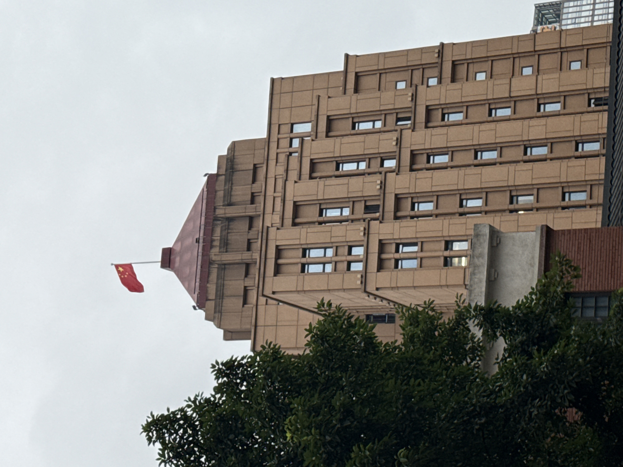 A tall, brown, multi-story building dominates the frame, with a Chinese flag flying from its peak. The building has a stepped, Art Deco-inspired architectural style, characterized by vertical and horizontal accents framing rectangular windows. In the foreground, a lush green tree partially obscures the lower left portion of the building. The sky is overcast and grey. The image appears to be taken from a low angle, looking upwards. The location is identified as Yuzhong District, China, which is known for its historical architecture and prominent landmarks.