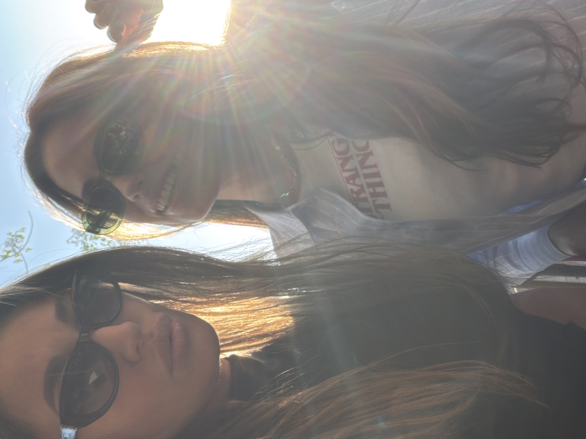 The image is a low-angle, sunlit photo of two women wearing sunglasses. The woman in the top portion of the frame is smiling and wearing a white T-shirt with red text that reads "STRANG THINGS" (possibly a reference to the TV show "Stranger Things"). The bottom woman has dark hair and sunglasses, and is looking up at the camera. The location may be Bar, Montenegro, given the image context. However, there are no location markers such as street signs or landmarks to definitively confirm the location. The bright sunlight creates significant lens flare.