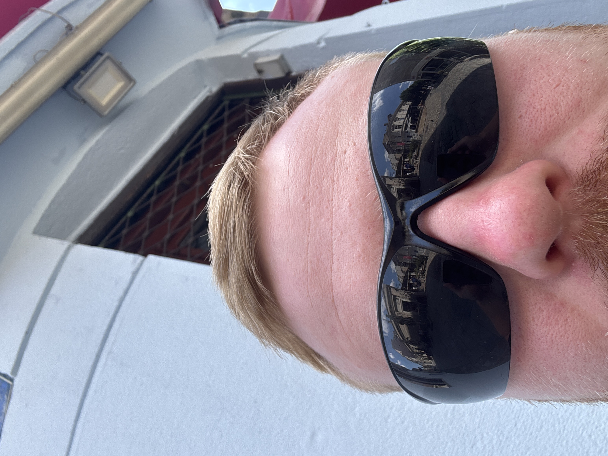 The image is a close-up, selfie-style shot focusing on the upper portion of a person's face, specifically their forehead, eyes obscured by black sunglasses, and nose. The sunglasses reflect a street scene with buildings and people. The background is a light blue wall with some architectural elements, possibly a light fixture. The person has light brown hair and stubble. The lighting suggests it is daytime. The reflection in the sunglasses shows a European-style street with old buildings, cobblestone pavement, and a few pedestrians.