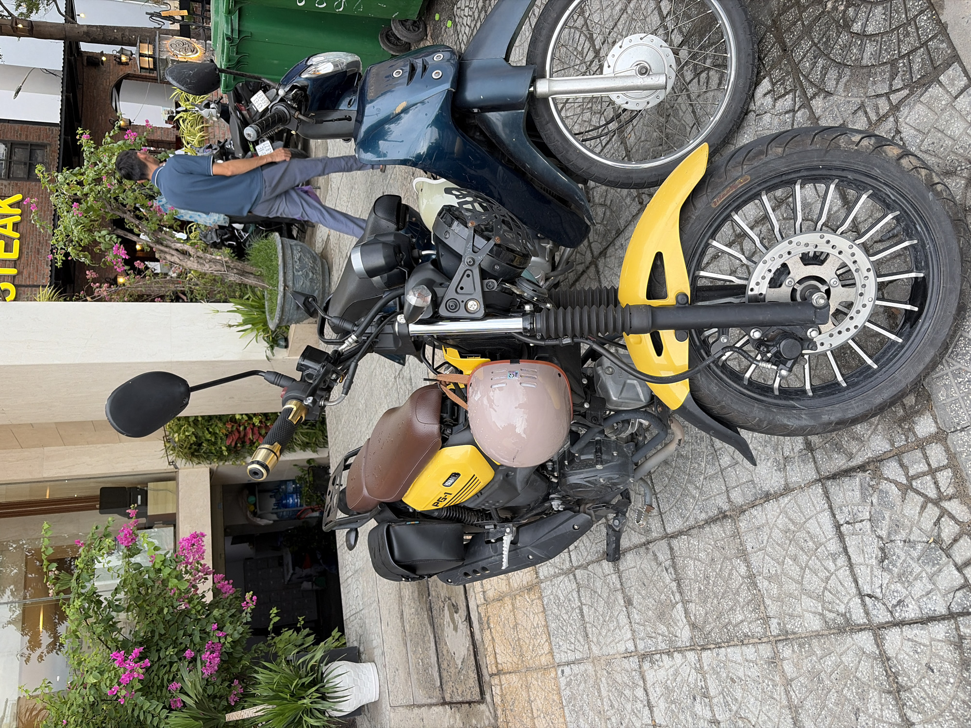 Two parked motorcycles are visible on a sidewalk in front of a building with a glass facade. A man in a blue polo shirt and gray pants walks away from the viewer, past the motorcycles and a large potted plant. One motorcycle is yellow and brown, with a pink helmet hanging from it. The other motorcycle is dark blue. The sidewalk is paved with square tiles. The background shows a building with plants and outdoor seating. The scene is likely in the city of Đà Nẵng, Vietnam, given the context of the request.