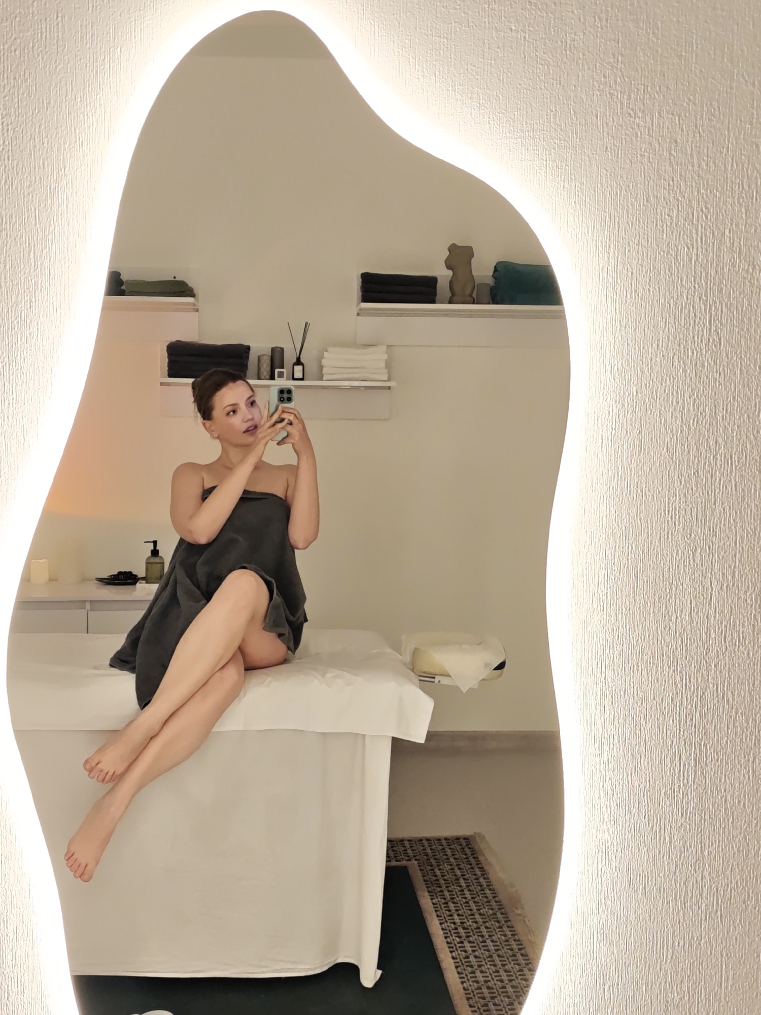 A fair-skinned woman with auburn hair sits on a white treatment table in what appears to be a spa or wellness center, as reflected in an illuminated, organically-shaped mirror. She is draped in a dark gray towel and holding a smartphone, seemingly taking a photo or video of herself.
Behind the woman, the wall features a minimalist design with stacked shelves holding folded towels, small bottles, and decorative items. Below the table, there is a visible area with a textured mat or rug. The wall on the right side of the mirror has a textured surface.
The location cannot be determined to be Vinnytsia, Ukraine, as there are no explicit clues in the image itself.