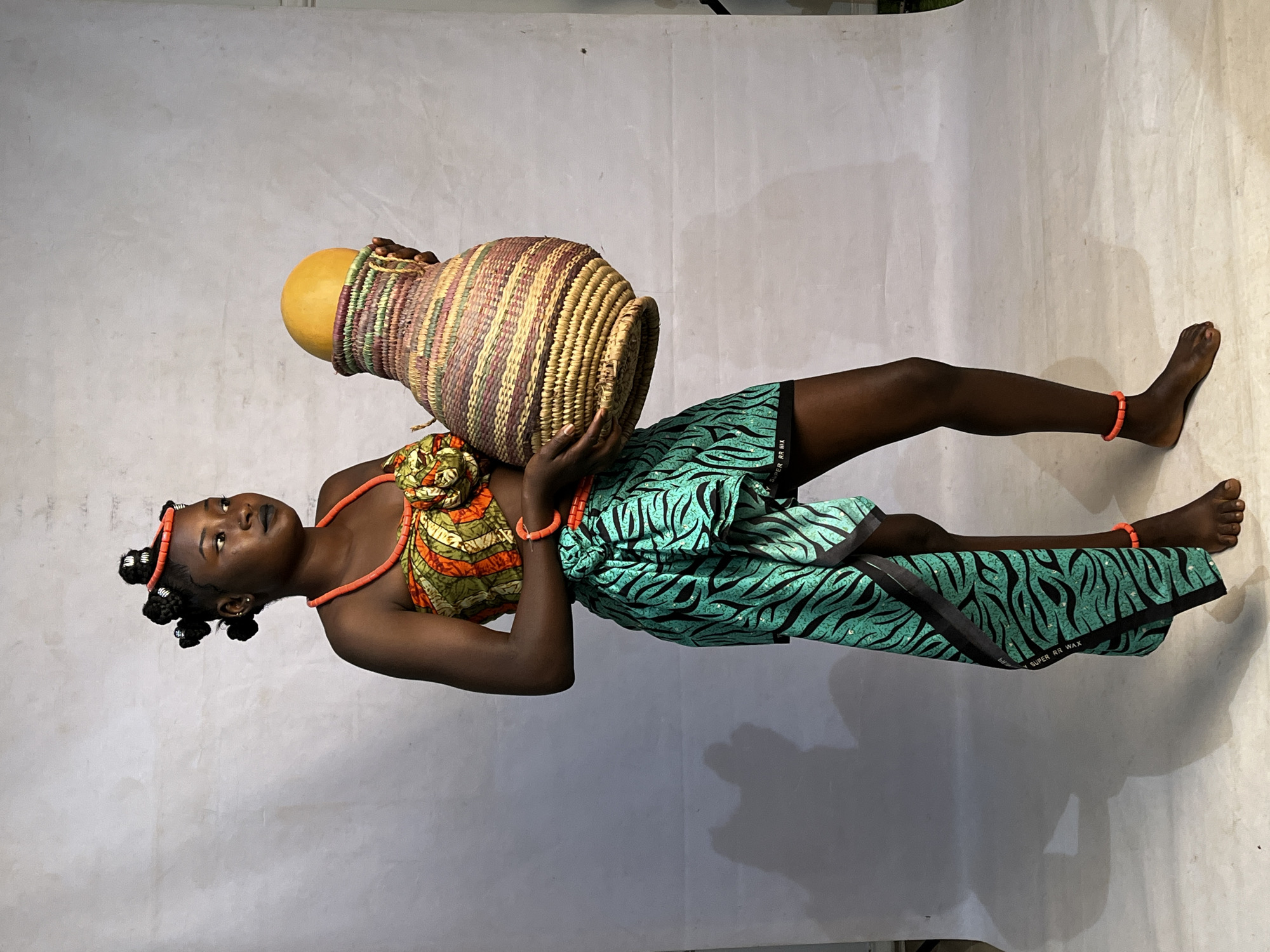A full-body, low-angle shot captures a young Nigerian woman standing against a plain, light gray backdrop. She is positioned on the left side of the frame, looking to her right. Her dark hair is styled in several intricate buns around her head, adorned with orange beads that also form a necklace and bracelets on her wrist and ankle. She wears a vibrant, patterned bandeau top with orange, green, and yellow hues, decorated with a floral element at the center. A flowing, teal skirt with a black tiger-stripe pattern is wrapped around her lower body, extending down her right leg. The woman is holding a large, intricately woven basket with both hands. The basket is filled with a bright yellow, spherical object. The lighting is soft, casting a shadow of the woman and the basket onto the backdrop. The image appears to be taken indoors, possibly in a studio, with no indication of the city of Barakin Akawo, Nigeria.