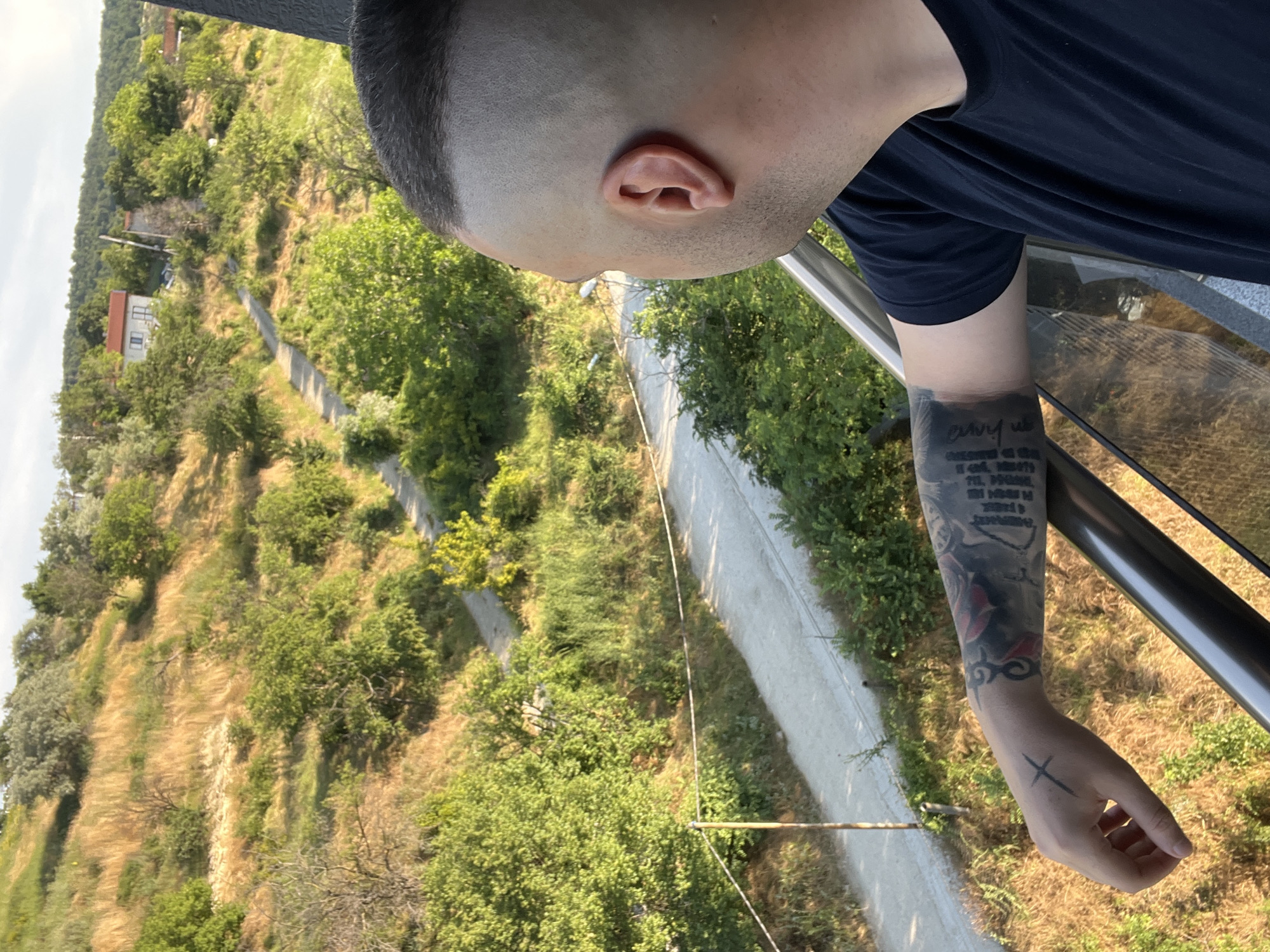 A man with a shaved head and tattoos on his arm stands on a balcony, looking out at a rural hillside. The hillside is covered in dry grass, green bushes, and scattered trees. A dirt road winds its way up the hill, with a few small houses visible in the distance. The sky is overcast. The man is wearing a dark t-shirt. The scene is likely located in the outskirts of Varna, Bulgaria, given the vegetation and architecture.