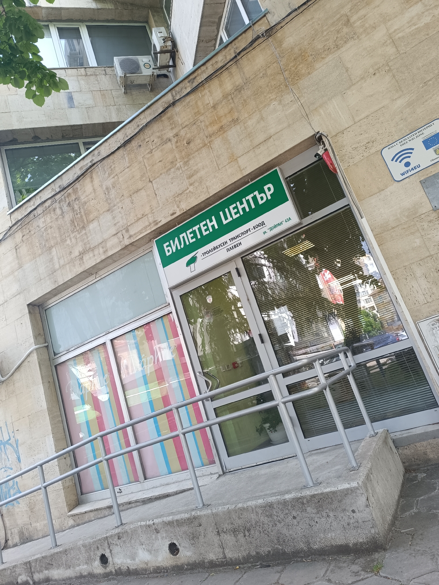 The image shows the entrance to a "Ticket Center" in Pleven, Bulgaria. The building is made of beige stone. Above the glass doors, a green sign displays "БИЛЕТЕН ЦЕНТЪР" (Ticket Center) in Bulgarian, along with "ТРОЛЕЙБУСЕН ТРАНСПОРТ • ЕООД ПЛЕВЕН" (Trolleybus Transport EOOD Pleven) and the address "ул. 'ДОЙРАН' 43А" (Doyran Street 43A). Colorful stripes decorate the window to the left. A gray metal handrail provides accessibility to the entrance, which has a concrete ramp. A sign indicating a free WiFi zone with the "WiFi4EU" logo is attached to the wall to the right of the entrance. The reflection in the glass doors shows a street scene with trees and buildings. An air conditioning unit is visible on the building facade above the ticket office.