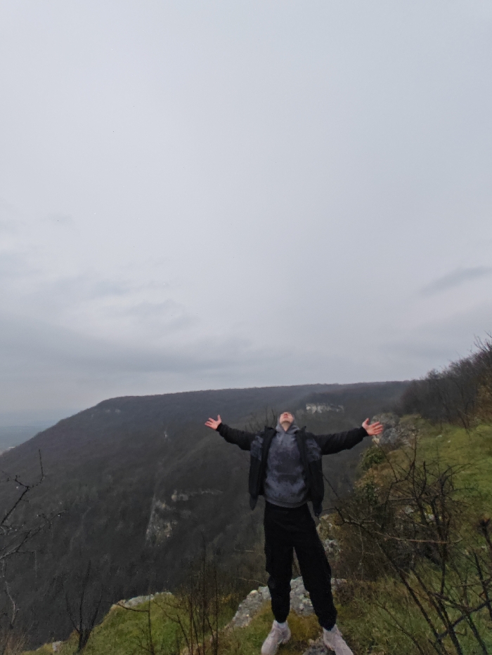 A male subject stands centrally on a rocky, grassy cliff edge overlooking a forested valley in the vicinity of Shumen, Bulgaria. The sky above is uniformly overcast and light grey, occupying the upper portion of the image. The individual is dressed in a black outer jacket over a grey and black patterned hooded sweatshirt, black track pants, and light-colored athletic shoes. He is positioned with his arms outstretched horizontally, palms facing upwards, and his head tilted back, looking towards the sky. The immediate foreground features sparse green vegetation and bare, thorny branches. In the mid-ground and background, dense deciduous forests cover rolling hills, extending towards a distant, faintly visible urban area on the left horizon, which is part of the city of Shumen.