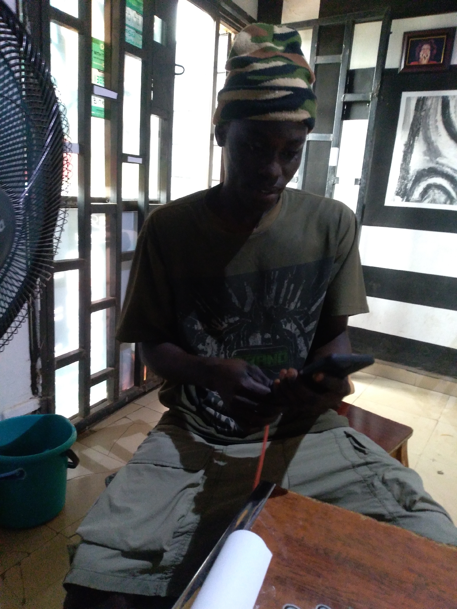 In Jos, Nigeria, a young Black male wearing a camouflage beanie and a dark green t-shirt is seated on a wooden chair. He is holding a smartphone in his hands, appearing to be engaged with it. The background reveals a room with large, divided windows that let in natural light. On the wall to the right, there are framed pieces of art, including a small portrait and a larger charcoal sketch. To the left of the frame, a black electric fan is visible. The floor is tiled, and a teal bucket sits on the floor to the left. A wooden table is in the foreground, partially obscuring the lower body of the individual.