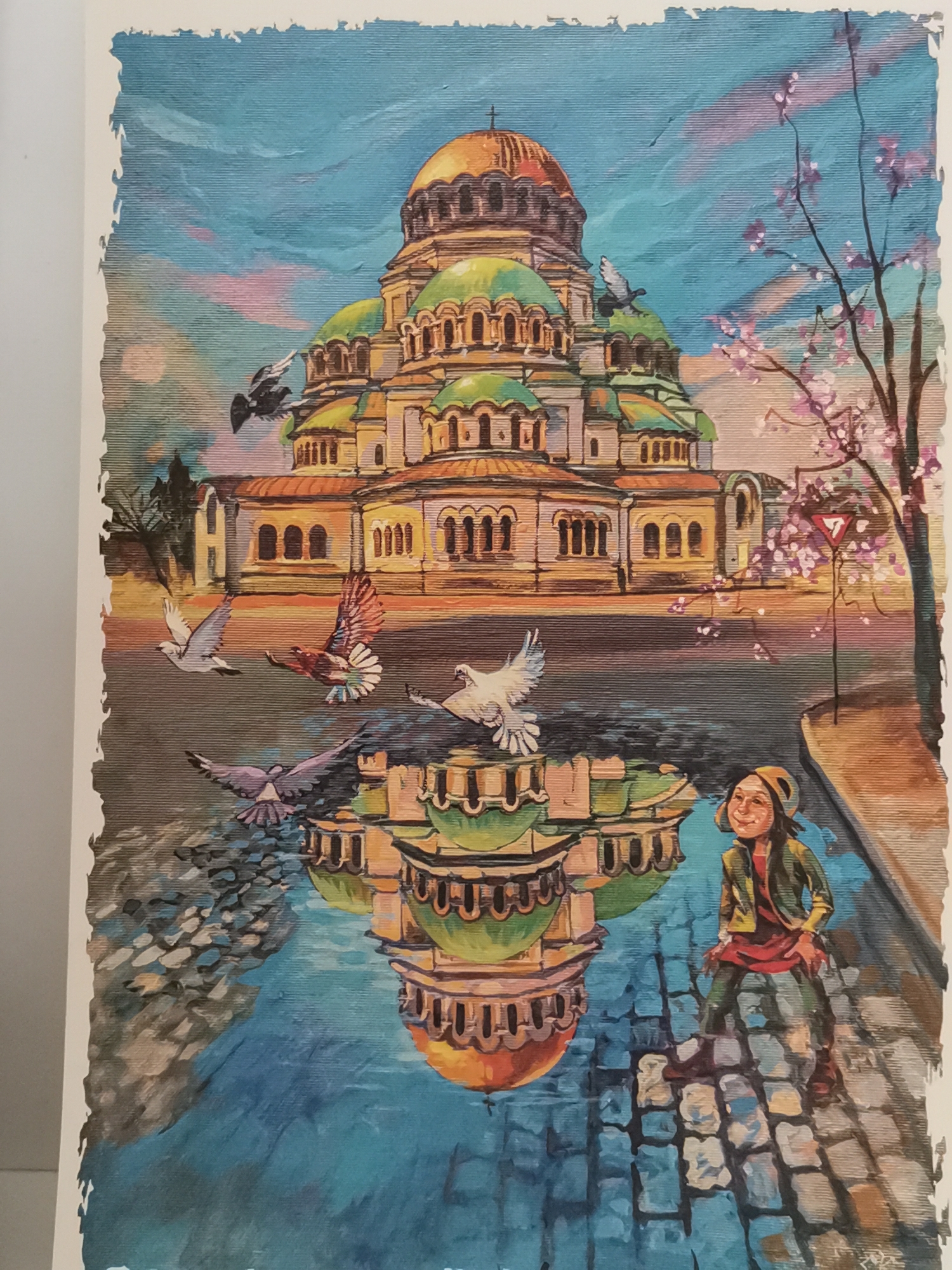 This detailed image depicts the Alexander Nevsky Cathedral in Sofia, Bulgaria, reflected in a large body of water on a paved surface. The prominent cathedral, characterized by its golden and green domes and light-colored walls, dominates the upper half of the composition, with its almost perfect reflection occupying the lower half. Six pigeons are in various stages of flight: one dark pigeon is high on the left, another dark pigeon appears to be landing on a green dome on the upper right, two white pigeons are flying low to the left of the reflected water, a reddish-brown pigeon with white tail feathers flies right above the water, and a purplish-gray pigeon flies diagonally downwards close to the water. On the right, a young person wearing a yellow beanie, a green jacket, and a red shirt stands on a cobblestone path, looking up with a slight smile towards the cathedral and pigeons. A tree with pink blossoms and a red triangular 'give way' traffic sign on a pole are also visible on the right. The sky, visible in the upper background, is blue with streaks of pink and purple clouds, suggesting dawn or dusk. The overall image has a painted, illustrative quality with visible texture.