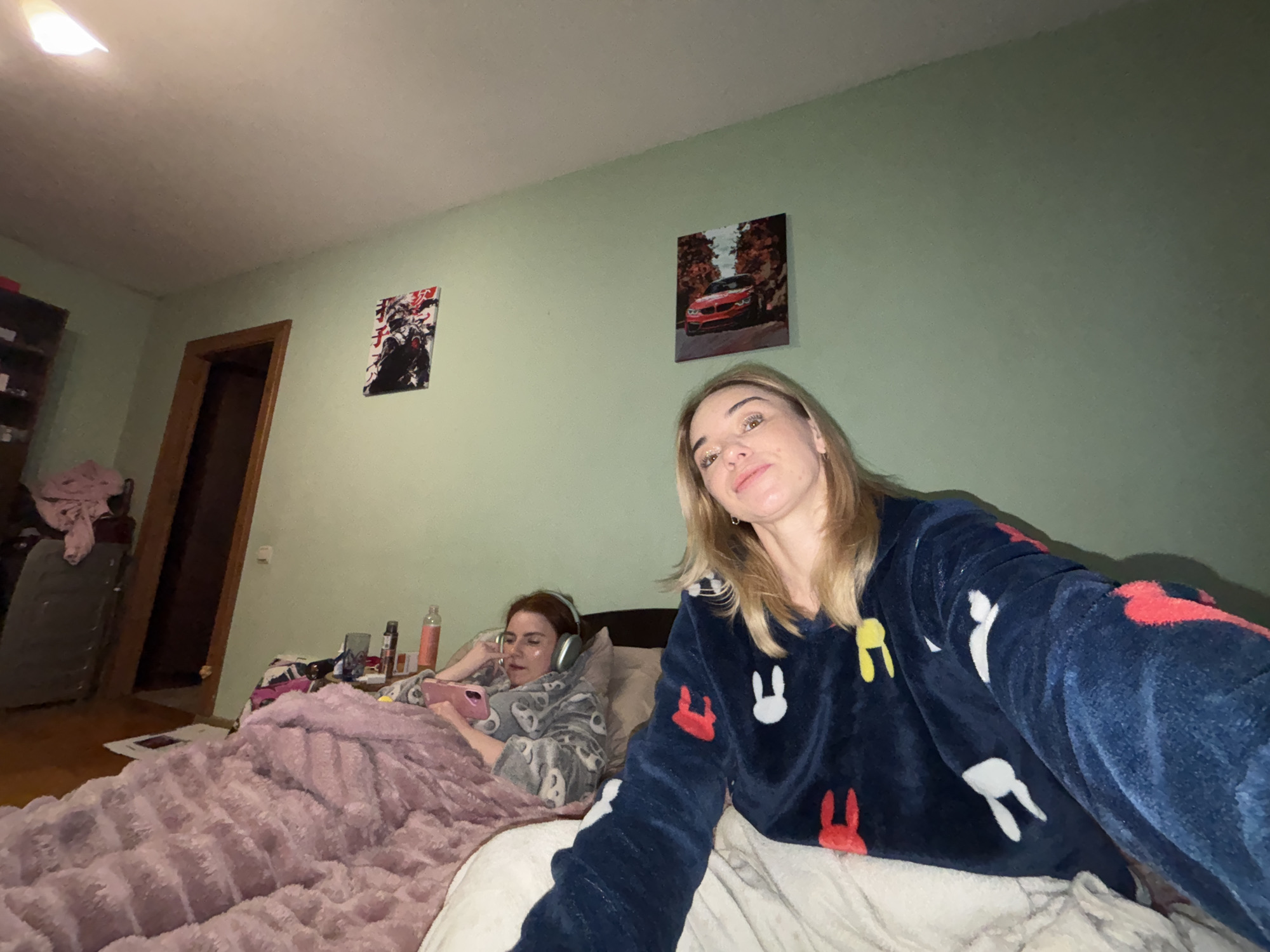 Two women are in a bedroom. One woman is closer to the camera, wearing a navy blue pajama top with white, red, and yellow bunny designs. She has fair skin and blonde hair. The other woman is lying in bed further away, wearing gray pajamas with white designs and headphones. She is using a pink mobile phone. The bed is covered with a light pink blanket and a white sheet. The room has light green walls. There are two framed pictures on the wall behind them: one of a motorcycle and one of a red car. A doorway can be seen on the left. A small table beside the bed has bottles and other items on it. An open closet is visible in the background. The location is Unknown, Unknown.