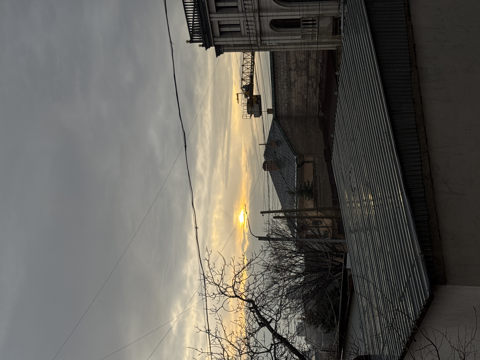 The image presents an urban landscape in Yerevan, Armenia, observed under a cloudy sky during either sunrise or sunset. The sun, appearing as a bright yellow-orange disc, is partially obscured by gray clouds near the horizon, serving as the primary light source and creating a backlighting effect.
In the immediate foreground, the dark, ribbed surface of a corrugated metal roof extends across the lower portion of the frame, angled downwards from left to right, with faint reflections of the sky visible on its ridges. Below this, a plain, light-colored wall is partially visible.
Bare, dark branches of a deciduous tree extend from the lower left, silhouetted against the bright horizon. A street light pole with an illuminated luminaire is positioned slightly to the right of the tree, its light appearing as a prominent yellow point. Multiple utility wires crisscross the upper and middle sections of the image.
In the midground, several structures are visible. A building with a dark, possibly tiled or slate roof and a visible chimney stands behind the street light. Adjacent to it, a wall with a rough, textured surface extends horizontally. On the far right, a multi-story building with classical architectural features, including a balustraded roofline, decorative cornices, and arched windows and balconies, is partially visible. The yellow jib of a tower crane extends horizontally behind the buildings on the right side of the frame. Distant, hazy building outlines are discernible on the far left horizon. No individuals or specific actions are depicted.