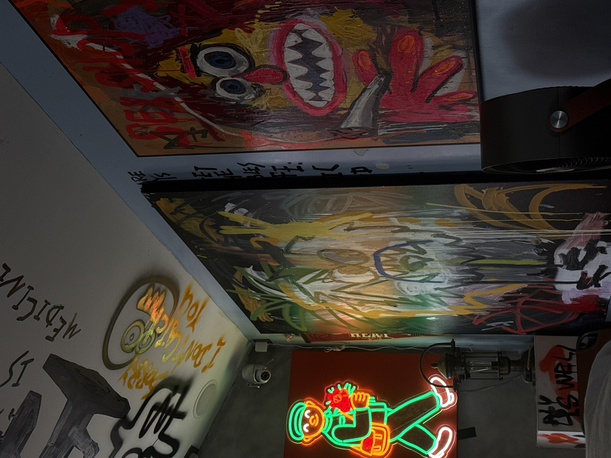 This media file captures an eclectic, art-filled interior space, likely a studio, cafe, or unique commercial establishment in Putuo District, China. The perspective is angled upwards, showcasing a ceiling adorned with two large, vibrant, graffiti-style paintings.
The upper painting features a cartoonish, expressive face with large eyes and a jagged, toothed mouth, set against a backdrop of red, yellow, and black brushstrokes. A red, multi-fingered hand is also visible. Partial text "SKULL" and some Asian characters, possibly "空," are integrated into the artwork. The lower ceiling painting is more abstract, with dynamic sweeps of yellow, green, black, white, and red. The word "REAL" is visible at its bottom center.
The walls are covered with various graffiti and text, including "MEDICINE" and phrases like "I DON'T KNOW" and "SORRY" in a metallic gold script within a circular emblem. Other black graffiti is partially visible. A prominent green and orange neon sign depicts a stylized figure, possibly a man in a hat, holding a bouquet of red flowers. Adjacent to the neon sign is an industrial-style light fixture with a caged bulb.
Other objects in the scene include a black, cylindrical fan or air purifier with a brown handle and a silver button, mounted near the ceiling. A white security camera is also visible on the wall. The lighting suggests an indoor evening or nighttime setting, with artificial light sources illuminating the artworks and creating a dynamic, urban ambiance. The overall impression is one of dense artistic expression and raw creativity.