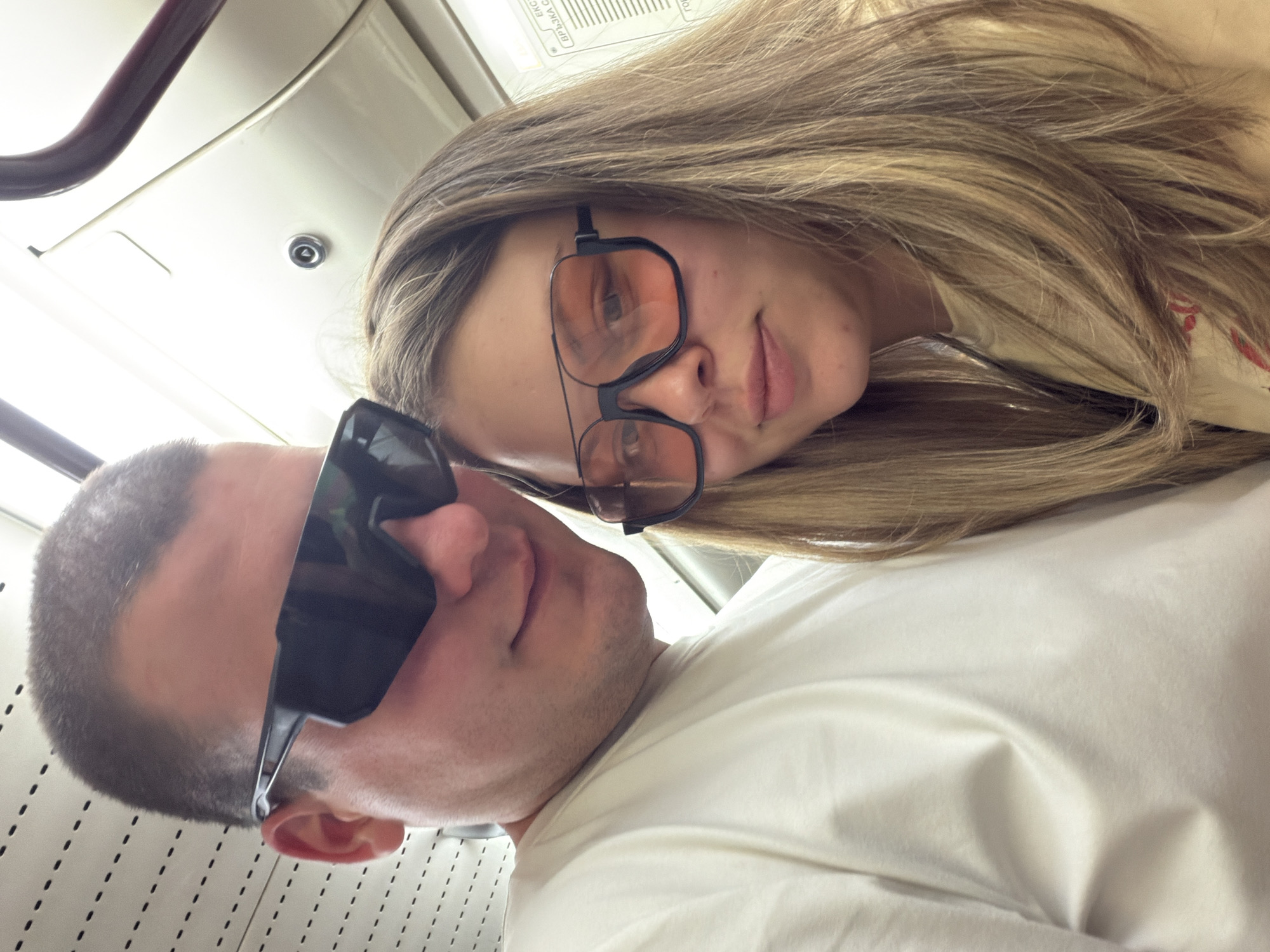 Two individuals, a male and a female, are posing for a selfie inside what appears to be a subway car, a common mode of public transport in Sofia, Bulgaria. The male, positioned on the left of the frame, is wearing a white t-shirt and large, black, angular sunglasses. The female, on the right, has blonde hair and is wearing orange-tinted, oversized square glasses and a light-colored top. The interior of the subway car is visible with a textured, off-white ceiling and a dark-colored handrail. A sign with Cyrillic text is partially visible in the background, further suggesting the location is within Sofia, Bulgaria. The lighting is bright, indicating the selfie was taken during the daytime.