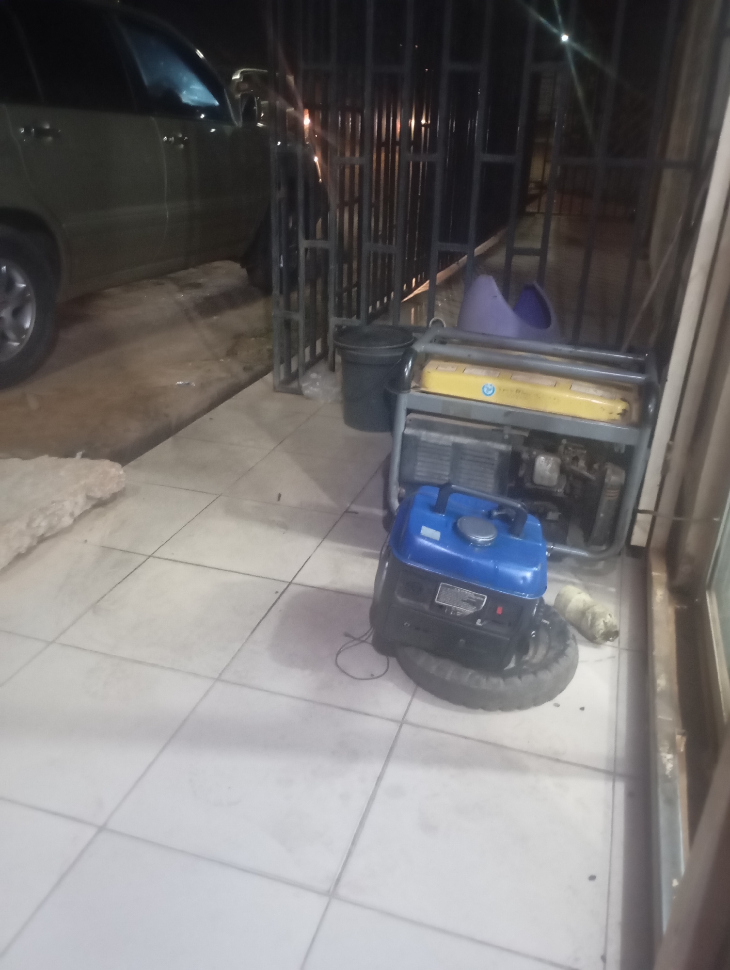 The image depicts an outdoor area at night, likely a residential or commercial premise in an urban or suburban setting. The foreground features a light-colored tiled patio or walkway, marked with dark grout lines. To the left, a dark-colored SUV (possibly olive green or dark grey) is partially visible, with its front right wheel and side panel facing the viewer. The vehicle's headlights are illuminated, casting a soft glow. It is parked on an elevated, darker, unpaved or concrete surface adjacent to the tiled area. A large, irregularly shaped piece of light-colored material, possibly insulation or rubble, rests on the unpaved ground near the tiled edge.
A dark, metal-framed security gate or fence stretches across the mid-ground, separating the vehicle area from the tiled space where several objects are placed. Beyond the gate, a bright light source is visible, suggesting a distant street light or an interior light from an unseen building.
On the tiled floor, in the right half of the image, are two portable power generators. The larger generator, further back, has a grey frame and a yellow fuel tank assembly with visible engine components. A smaller, blue-colored generator is positioned in front of it, resting on a black, deflated tire or rubber mat. This smaller generator has a silver fuel cap and a black control panel. To the right of the larger generator, a small, white, cylindrical object (possibly a rolled towel or piece of pipe) lies on the tiles. Behind the larger generator, against the fence, a dark bucket and a purple plastic object (resembling a child's potty training seat or a small basin) are visible. To the far right, a vertical wooden or metal structural element of an adjacent building or entrance frame is partially seen. No individuals are present, and no actions are currently taking place.