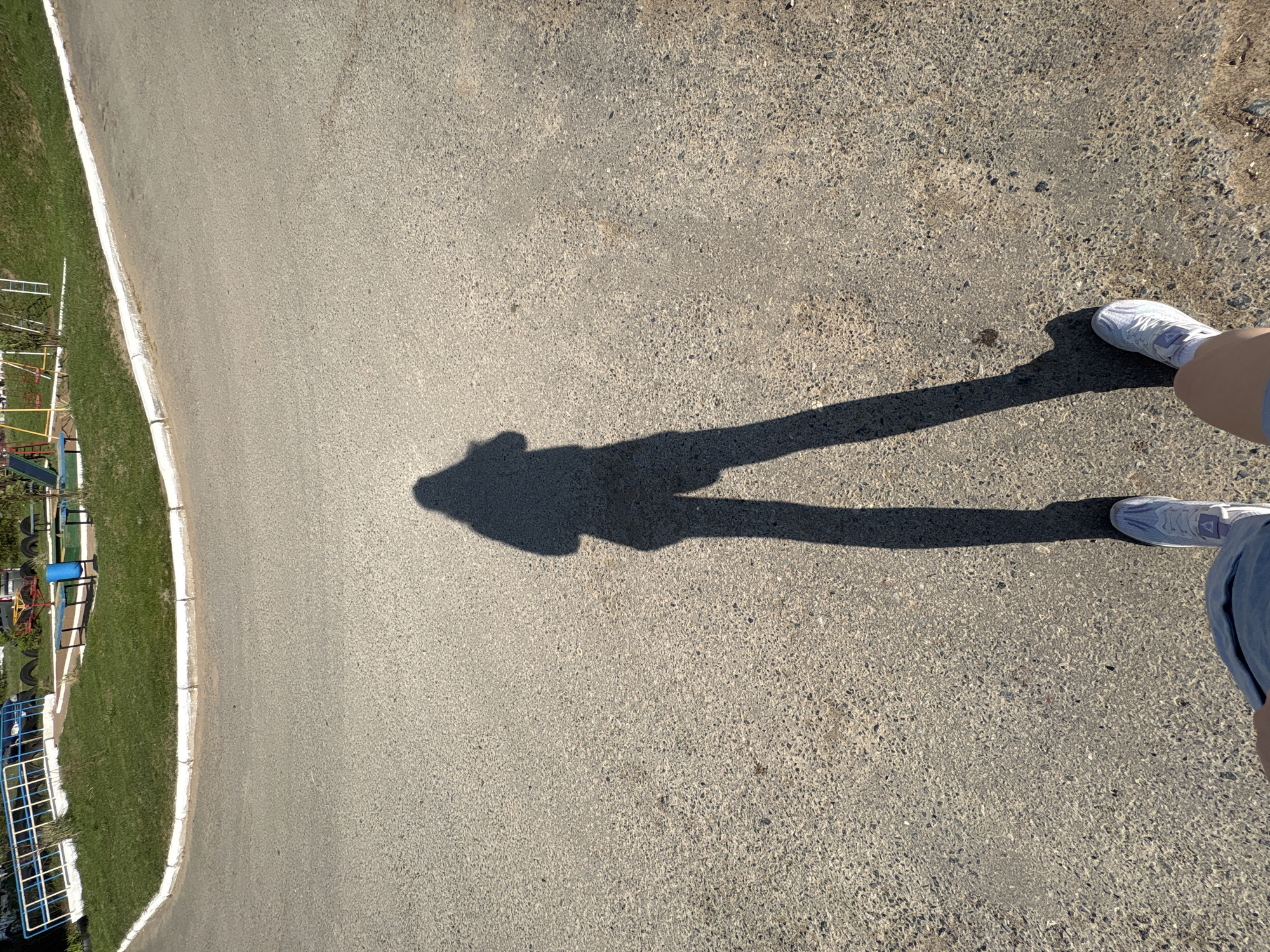A street scene is captured from an elevated perspective, potentially from the vantage point of a person looking down at the ground. The primary surfaces are paved, appearing to be either a road or a sidewalk. A shadow of a person, complete with identifiable legs and feet wearing athletic shoes, stretches across the paved surface.
To the left of the pavement is a grassy area with playground equipment visible. This suggests the image was taken near a park or recreation area.
Based on the location of the city of Chaikovsky, Russia, the environment likely experiences seasonal variations in weather and sunlight, although the conditions depicted in the image appear to be of a bright, sunny day. The image does not contain any additional information as to who or what captured this picture.
