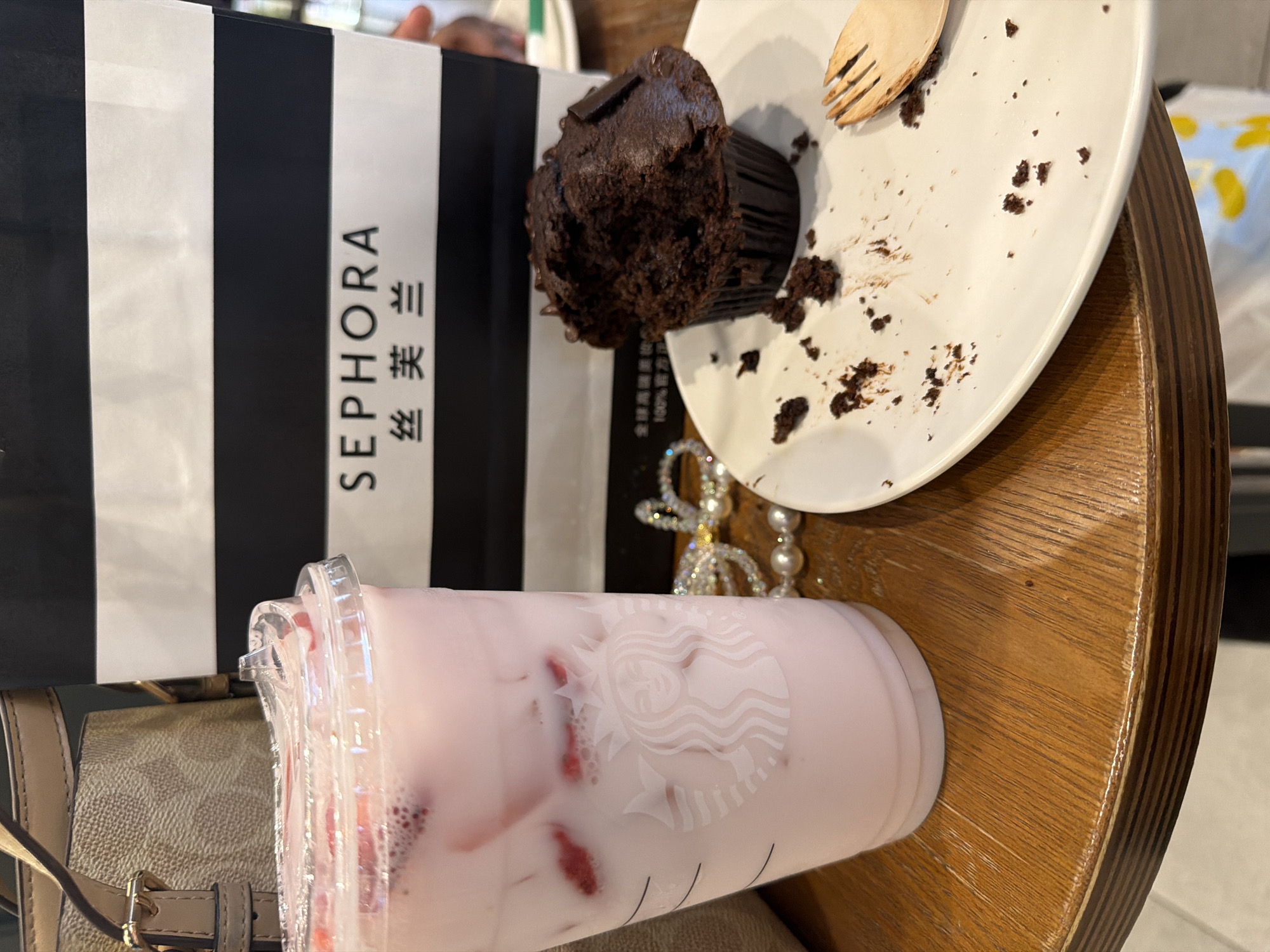 An overhead shot captures a collection of items on a wooden table, suggesting a casual break or outing. The scene features a partially eaten chocolate muffin, a pink beverage in a Starbucks cup, a Sephora shopping bag, and a beige purse.
The dark chocolate muffin, still in its paper liner, rests on a white ceramic plate. A portion of the muffin has been eaten, leaving crumbs scattered across the plate, alongside a small, light-colored wooden fork. Adjacent to this, a clear plastic Starbucks cup contains a light pink, opaque drink with ice and visible red fruit pieces, likely strawberries. The iconic Starbucks siren logo is clearly visible on the cup's surface.
Behind these items stands a black and white striped Sephora shopping bag. The brand name "SEPHORA" is printed vertically in capital letters on a white stripe, immediately followed by the Chinese characters "丝芙兰" (Sīfúlán), the brand's name in Chinese. To the left, a beige or light brown purse with a patterned texture and a gold-toned buckle is partially visible. A piece of shimmering jewelry, possibly a pearl or rhinestone necklace, is also visible on the table.
The setting appears to be indoors, with a wooden tabletop featuring a distinct grain. The presence of the Sephora bag with Chinese text strongly indicates the location is in China, aligning with the provided context of Yuhuatai District. The image portrays a moment of relaxation, possibly after a shopping trip.