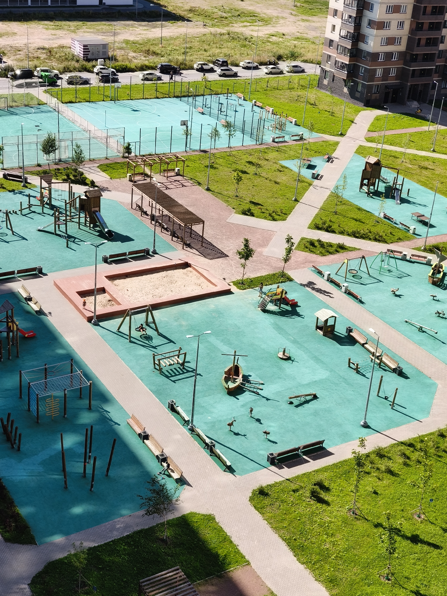 The aerial photograph, taken in Murino, Russia, depicts a modern, multi-functional urban recreational area. The scene is dominated by a bright, turquoise-colored rubberized surface designed for playgrounds and sports courts. Multiple distinct zones are visible, including two marked sports courts, likely for basketball or volleyball, enclosed by green fencing. Adjacent to these courts are open grassy areas and a designated parking lot with several cars.
The primary focus is on a large, well-equipped children's playground. This area features a variety of play structures made of wood and metal, including slides, swings, climbing frames, and a sandbox. Two wooden pergolas with benches provide shaded seating areas. Further into the complex, additional playground equipment is scattered across the turquoise surface, including seesaws, spinning toys, and a ship-shaped play structure. A series of wooden posts, possibly for a balance course, are also present.
Pathways constructed from light-colored paving stones wind through the recreational area, connecting different zones and leading to the surrounding buildings. These pathways are bordered by meticulously maintained green lawns and young trees, indicating a recent urban development. In the background, a tall, multi-story residential building with a modern facade stands prominently, casting long shadows across the scene. The overall impression is one of a thoughtfully designed, clean, and vibrant community space.