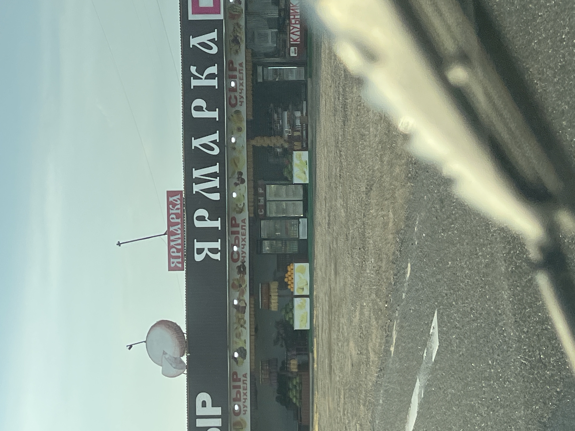 The image captures a market stall in Psekups, Russia, under a clear sky. The stall, primarily displaying agricultural products, features a large sign with the word "ЯРМАРКА" (Yarmarka, meaning fair or market) in white Cyrillic letters against a dark background. To the left of this sign, a stylized graphic of a sliced apple adorns the structure. Below the main signage, a banner displays smaller text: "СЫР" (Syr, meaning cheese) and "ЧУЧХЕЛА" (Chuchkhela, a Georgian candy made from nuts and grape juice). The stall itself is stocked with various items, including watermelons, oranges, and what appear to be other fruits and produce. Refrigerated display cases and shelving units are visible inside the stall. To the far right, a sign reads "КЛУБНИК" (Klubnik, meaning strawberry). The ground in front of the stall is unpaved, appearing to be a dirt or gravel surface. In the foreground, a portion of a vehicle's windshield and wipers is visible, suggesting the image was taken from inside a car.