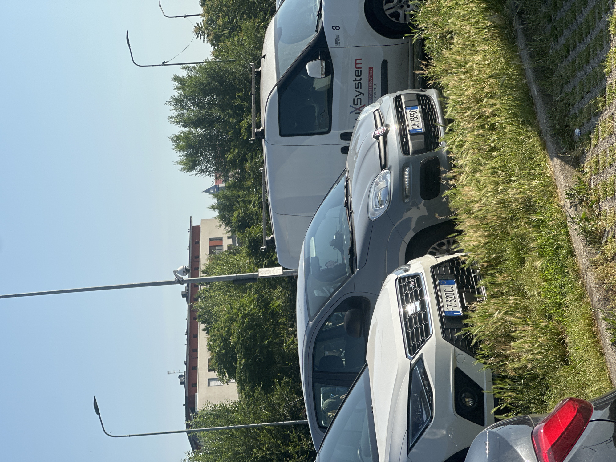 The image shows several vehicles parked in a parking area, with tall grass growing beside the parking spaces. A white van with the text "xSystem" and the number "8" is parked next to a gray Fiat Panda, which has the license plate "CA 735RC". In front of the Panda, a white car with the license plate "FZ-320CJ" is visible. In the background, there is a multi-story building and trees under a clear sky. Two street lights can be seen extending above the trees. Although the location is not explicitly mentioned in the image, the context clues suggest that it is a typical urban scene in an Italian city.