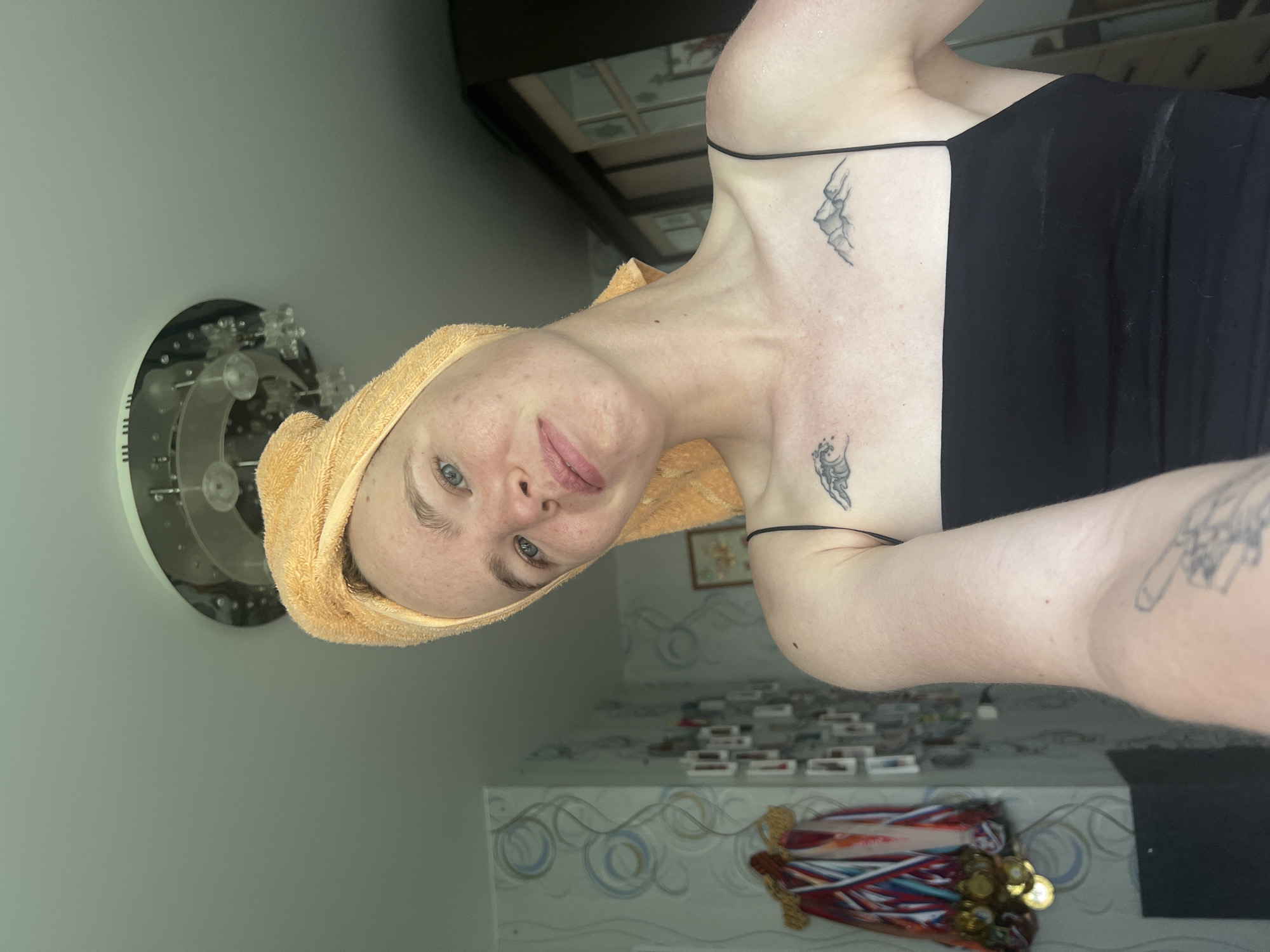A young woman with a towel wrapped around her head poses for a selfie indoors, likely in her home, in Chaikovsky, Russia. She is wearing a black tank top and has two small tattoos on her chest: a wave on the left and mountains on the right. The background shows a wall with patterned wallpaper and a collection of framed pictures and medals hanging on it. A large wardrobe is visible on the right side of the frame, and a ceiling light fixture is present above. The lighting suggests it is daytime.