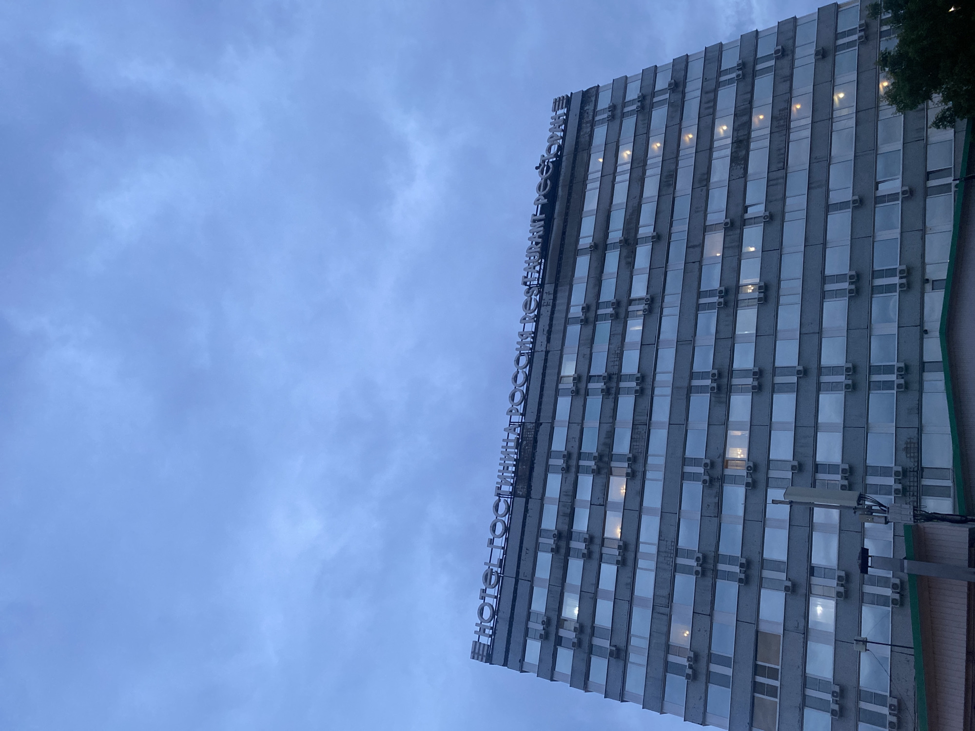 The image shows a building with a sign that reads "HOTEL KOSMOS" in large, silver, capital letters. The building is tall and appears to be made of concrete and glass, with many windows reflecting the cloudy sky. Several air conditioning units are visible mounted outside the windows. The sky is overcast and a pale blue, suggesting it might be daytime but not bright sunlight. The angle of the photo is from below, looking up at the building. A tree is partially visible on the right side of the frame. The building appears to be located in an urban or semi-urban setting. The overall mood is somewhat subdued due to the cloudy weather and the utilitarian architecture of the building. There is no activity of people visible in the image. The time of day appears to be evening or late afternoon. The setting is Novokuybyshevsk, Russia.